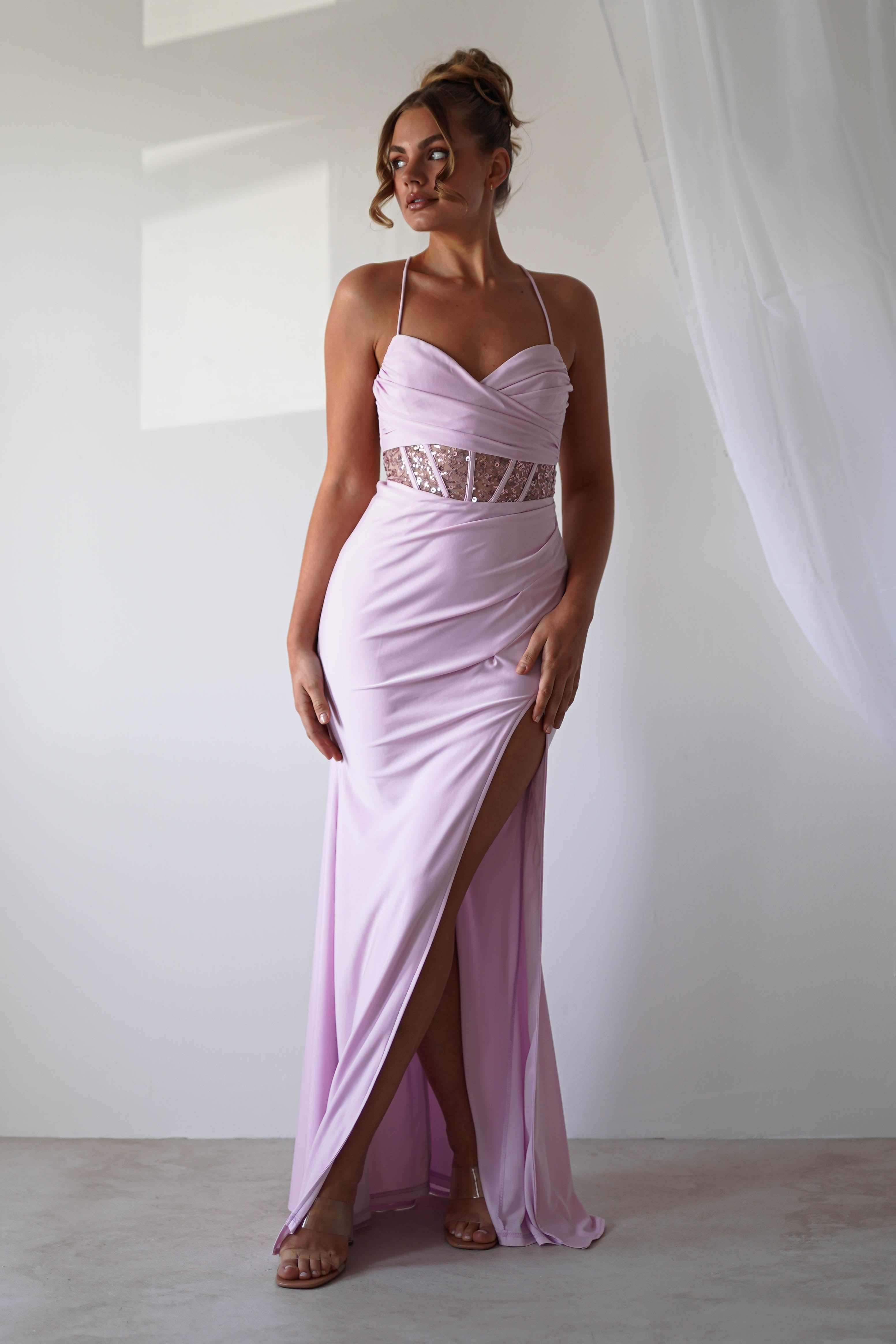 A woman in a light purple halter-neck gown with lace detailing and sequin accents poses elegantly against a white wall. The dress, perfect for formal events, features a thigh-high slit. Her hair is styled in an updo, and she wears strappy sandals beside a sheer curtain.