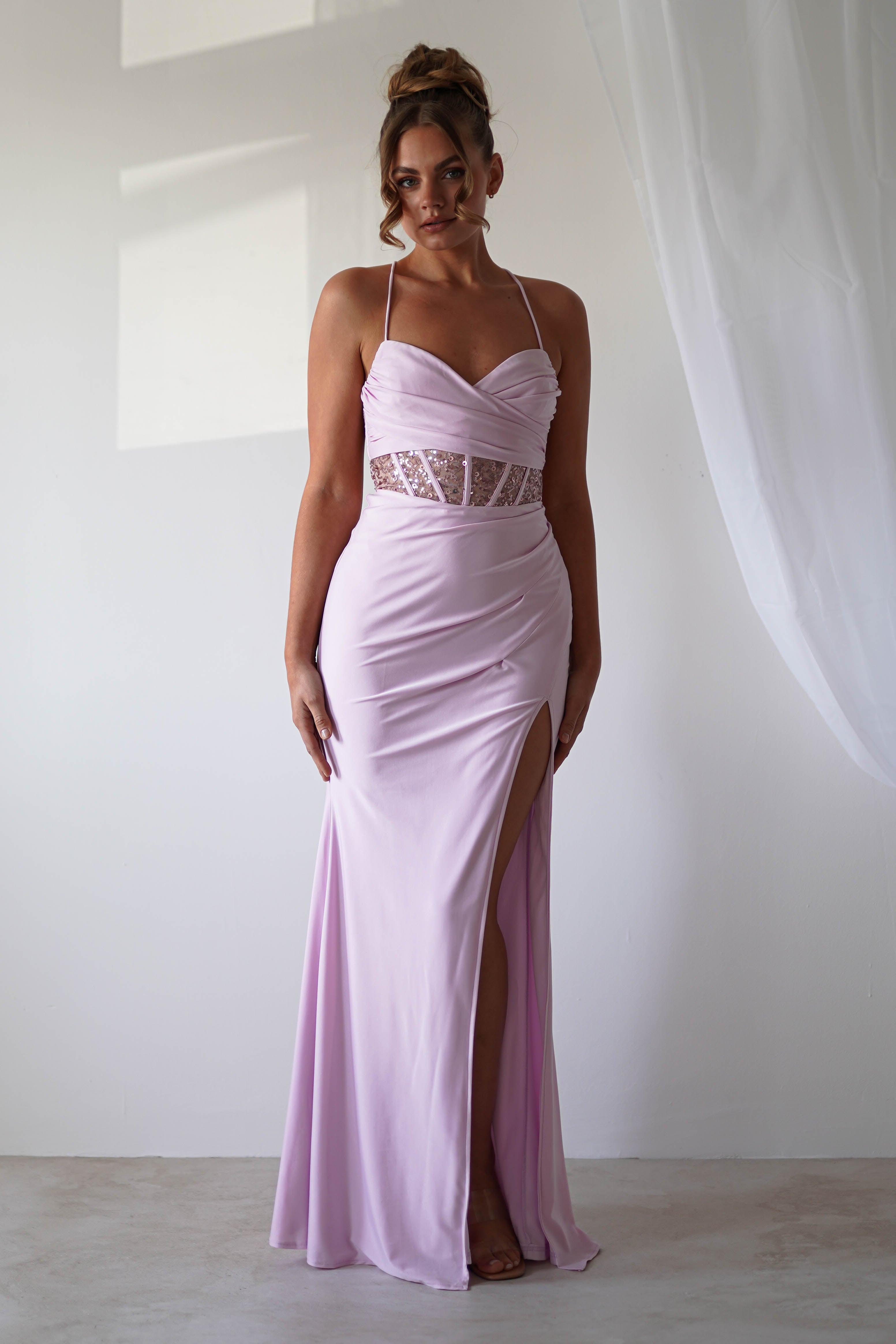A woman in a lavender gown with a halter neckline and sequin detail at the waist stands elegantly in a softly lit room with sheer white curtains. Her hair is styled in an updo, perfect for formal events, as the thigh-high slit adds an alluring touch.