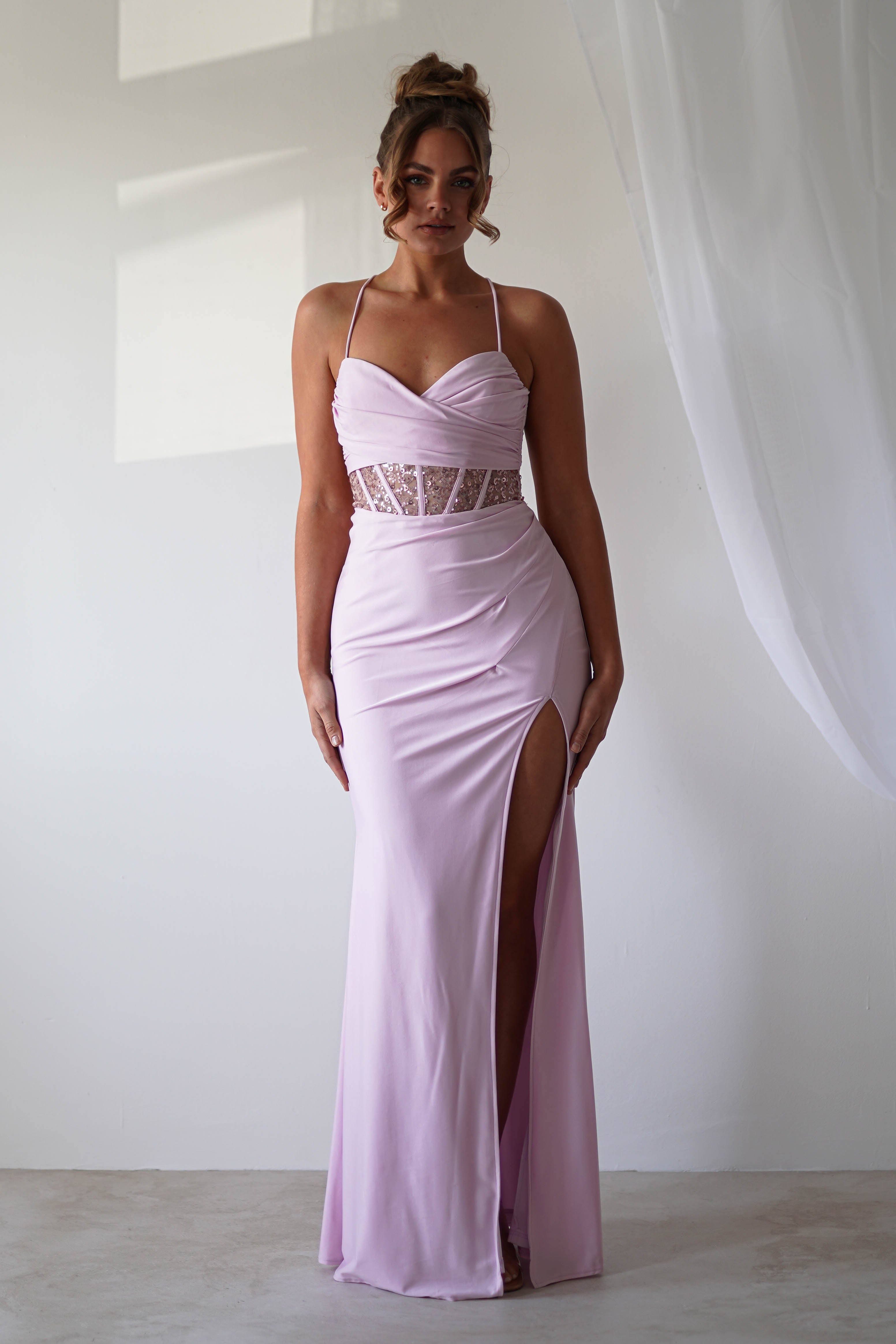 Woman in light pink, sleeveless, floor-length gown with lace and sequin detail stands against a minimalist background. Her hair is styled in an updo, and a sheer white curtain hangs to the side—a perfect look for formal events.