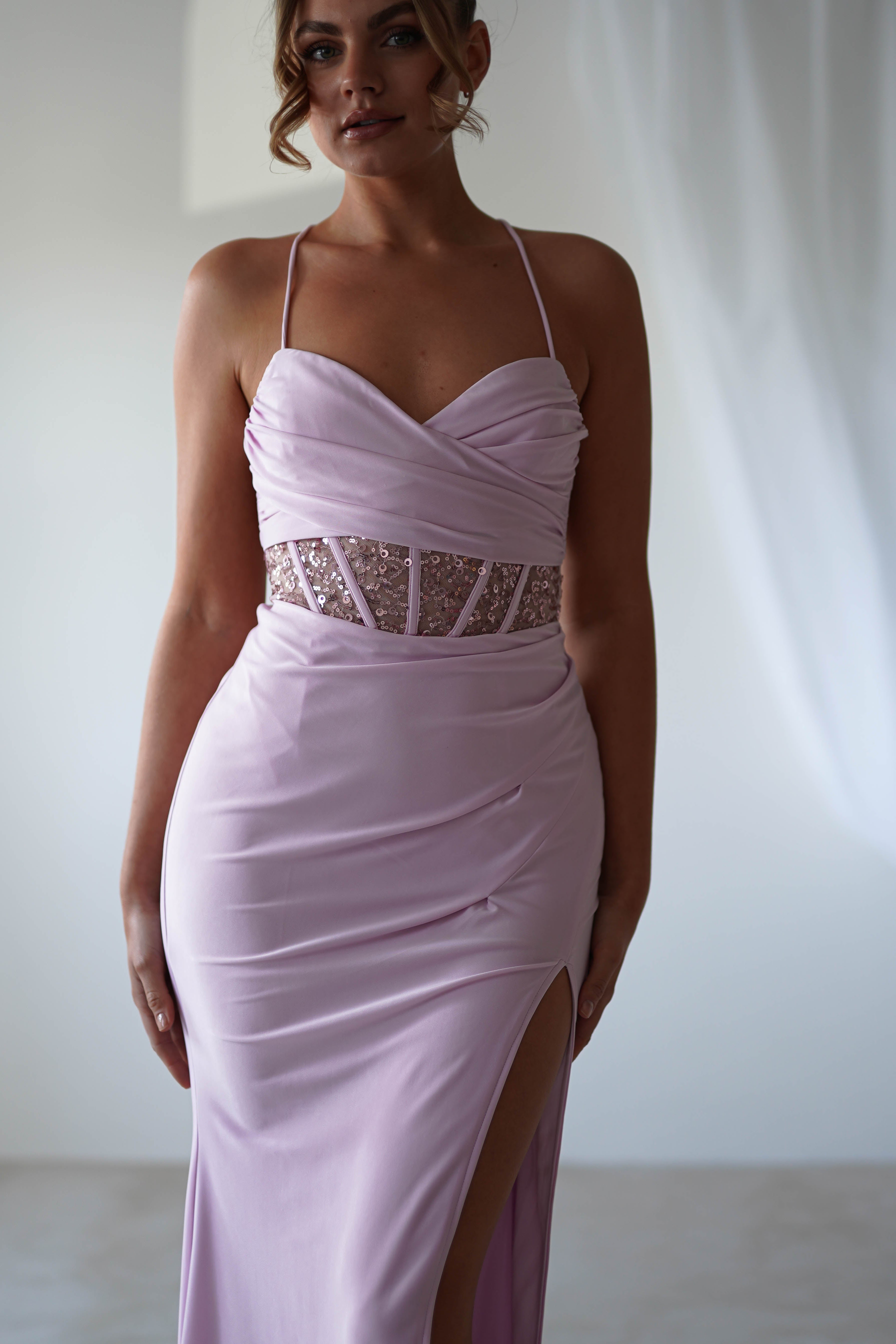 A person is wearing a lavender gown with thin straps and lace detailing around the midriff. The dress features a sweetheart neckline, sequin detail, and a thigh-high slit, making it perfect for formal events. The background is softly lit and neutral.