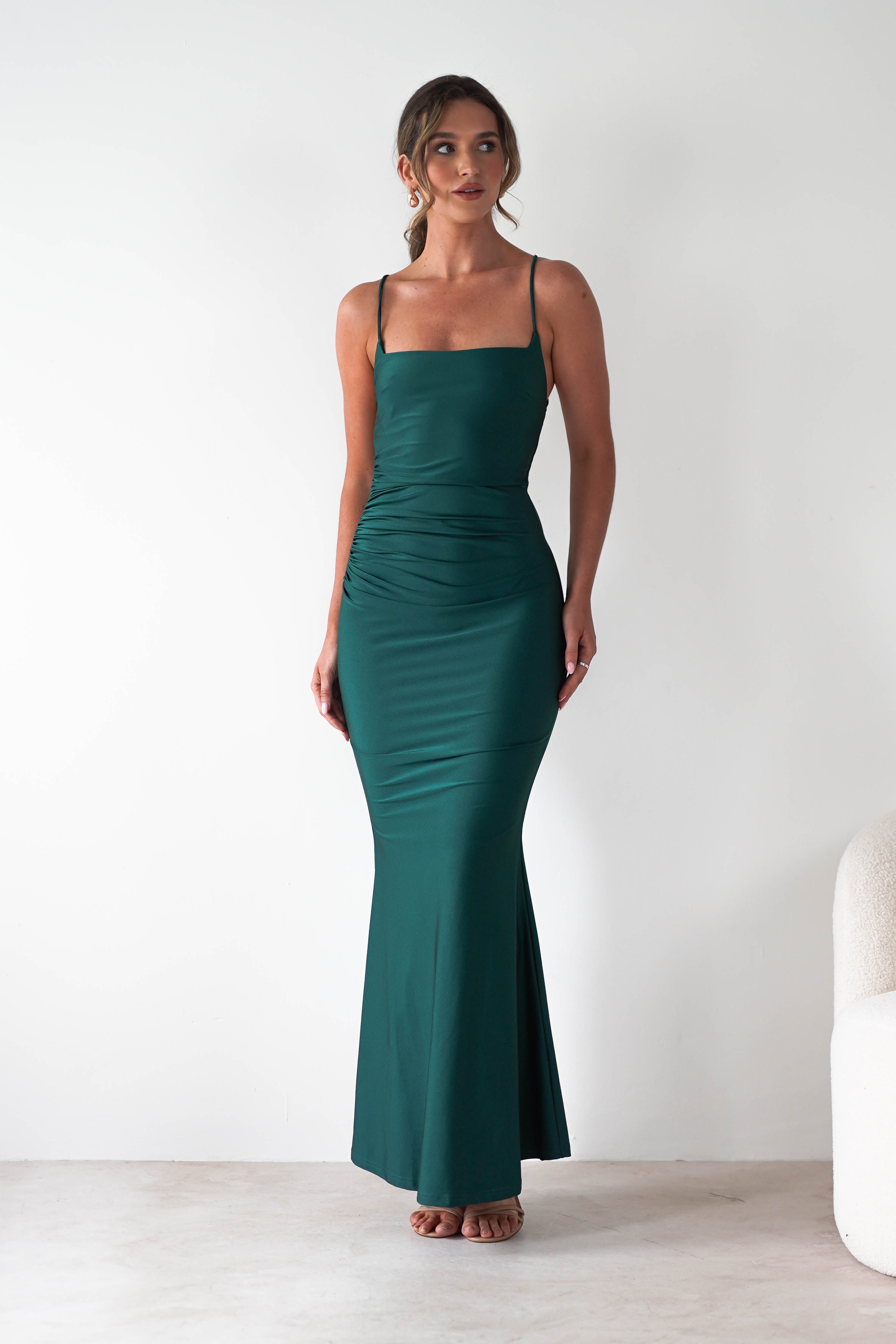 A woman stands indoors wearing a fitted, sleeveless green gown with thin straps and a square neckline. Perfect for formal occasions, the dress features subtle ruching at the waist and a flowing mermaid-style skirt against a plain white background.
