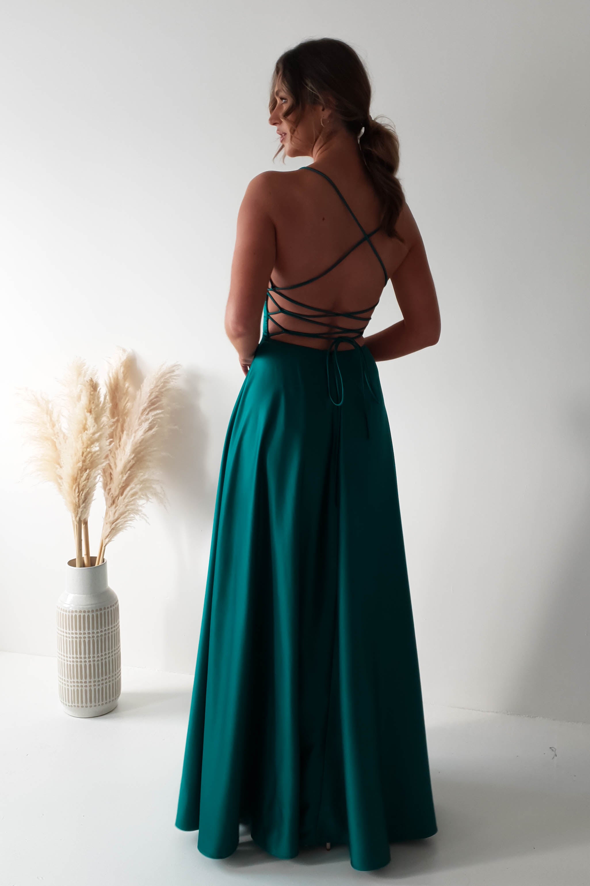 A woman stands with her back to the camera, wearing a teal, backless gown featuring crisscross straps and a daring high slit. She is in a minimalist room with a white wall and a vase of pampas grass on the floor.