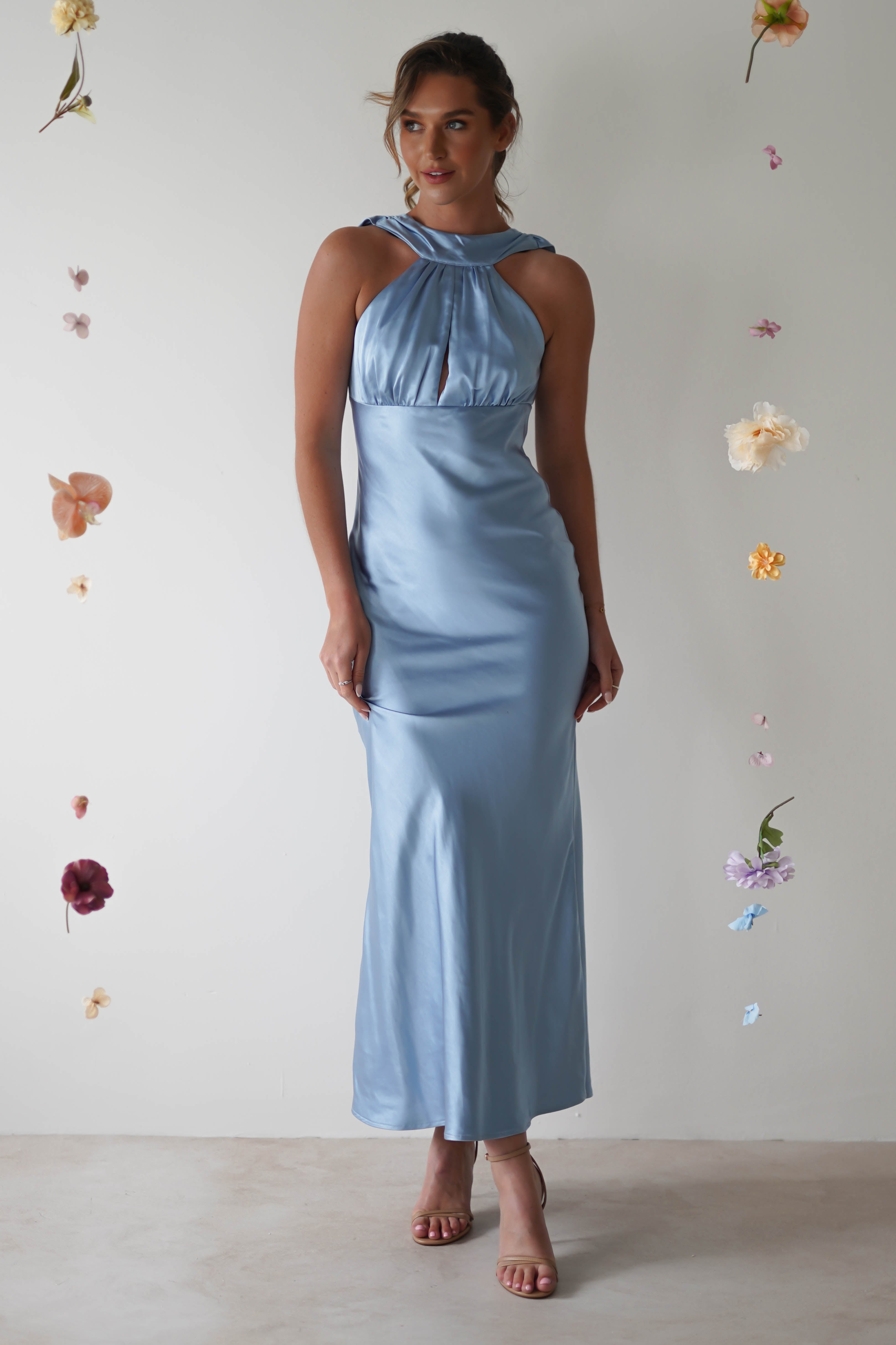 A woman wearing a light blue halterneck maxi dress poses against a plain backdrop. The satin material of the open-back gown flows elegantly to the floor, creating a fitted silhouette. Small, hanging flowers surround her, adding a whimsical touch to the scene.