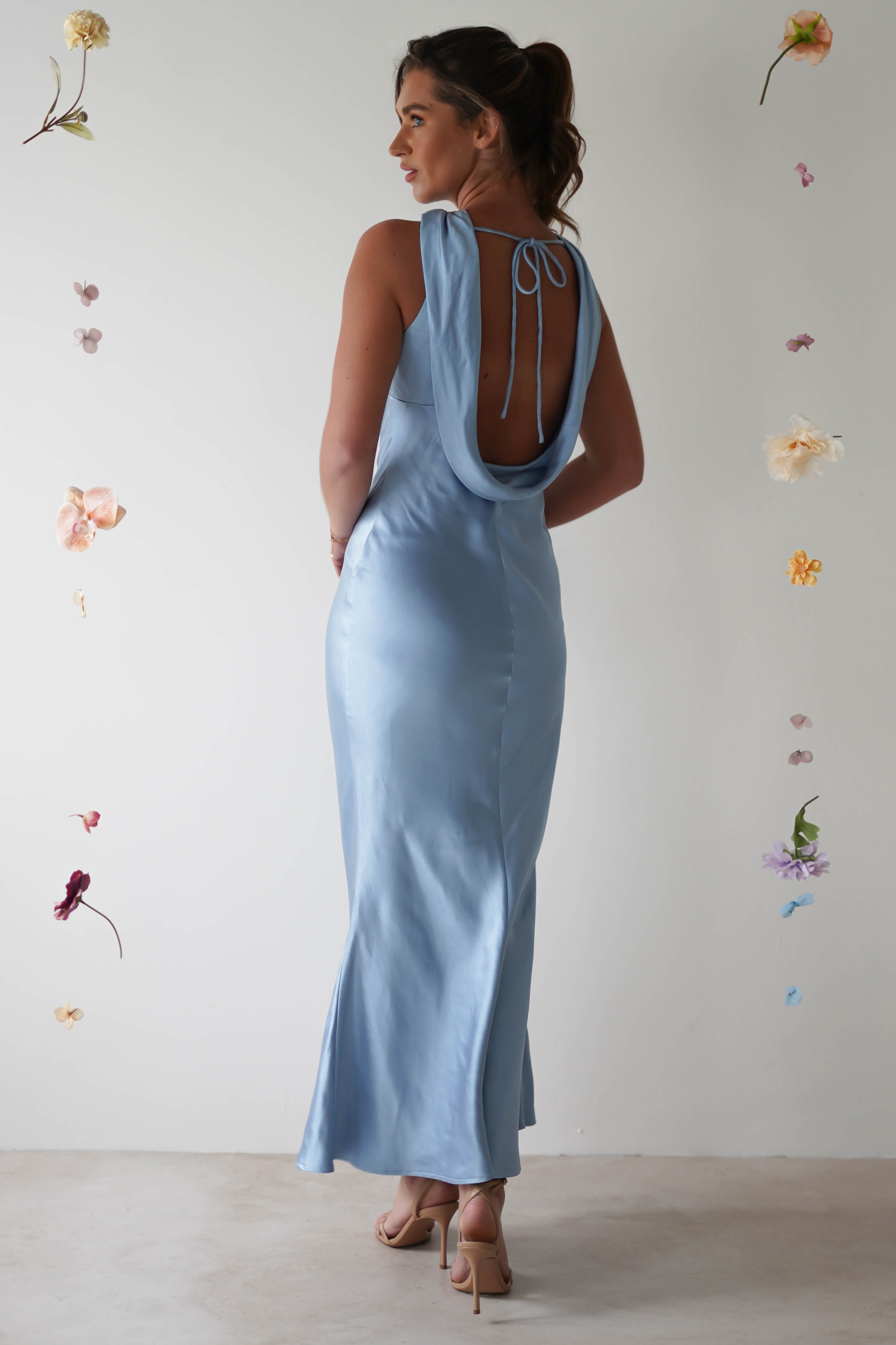 A woman in a light blue halterneck satin maxi dress with an open-back detail stands against a plain background, surrounded by suspended flowers. Her hair is elegantly pulled into a ponytail as she is viewed from behind.