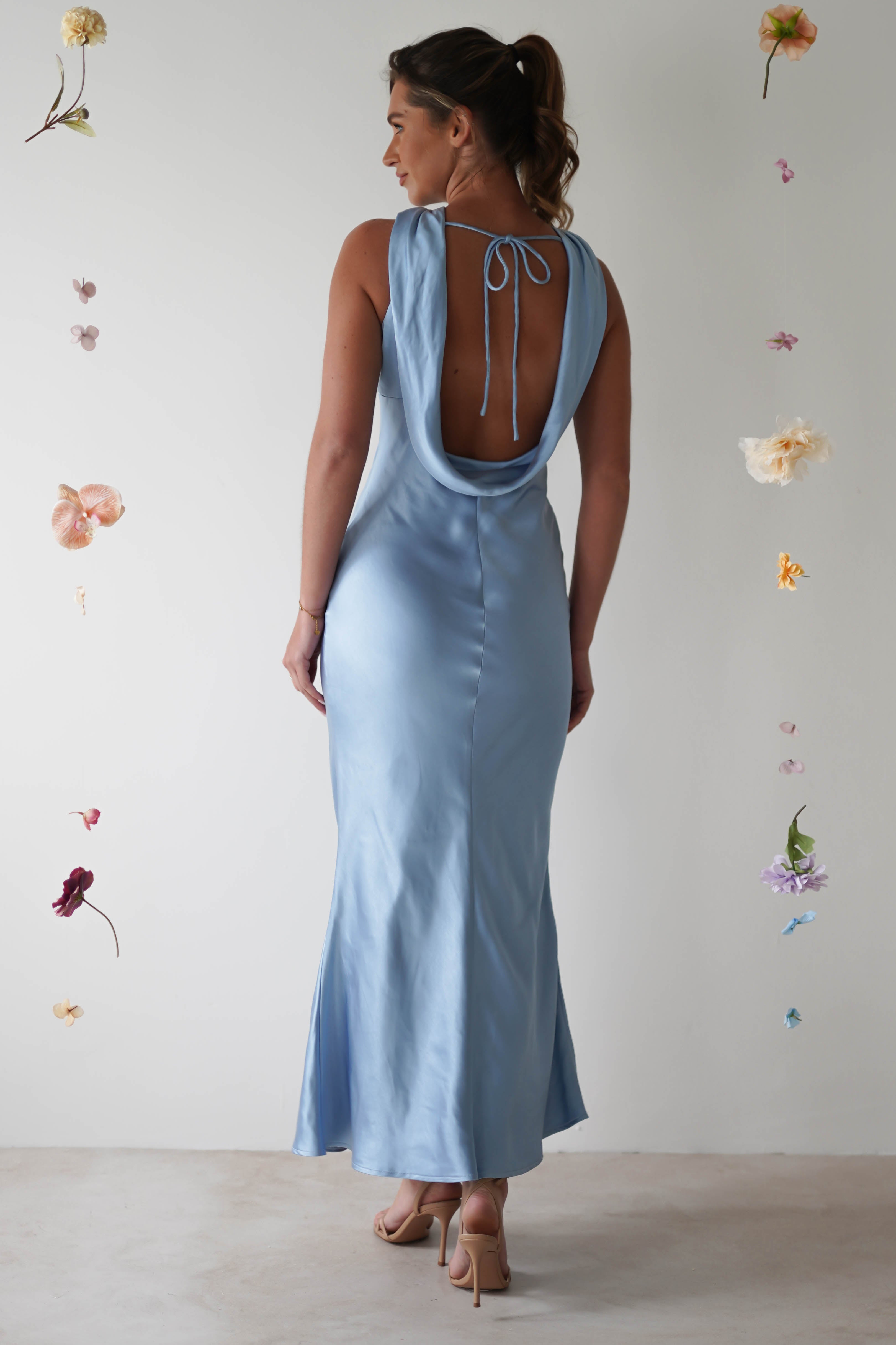A woman in a silky, light blue halterneck maxi dress stands with her back to the camera. Her hair is in a ponytail. The satin material shimmers as shes surrounded by floating, colorful flowers against a plain background.