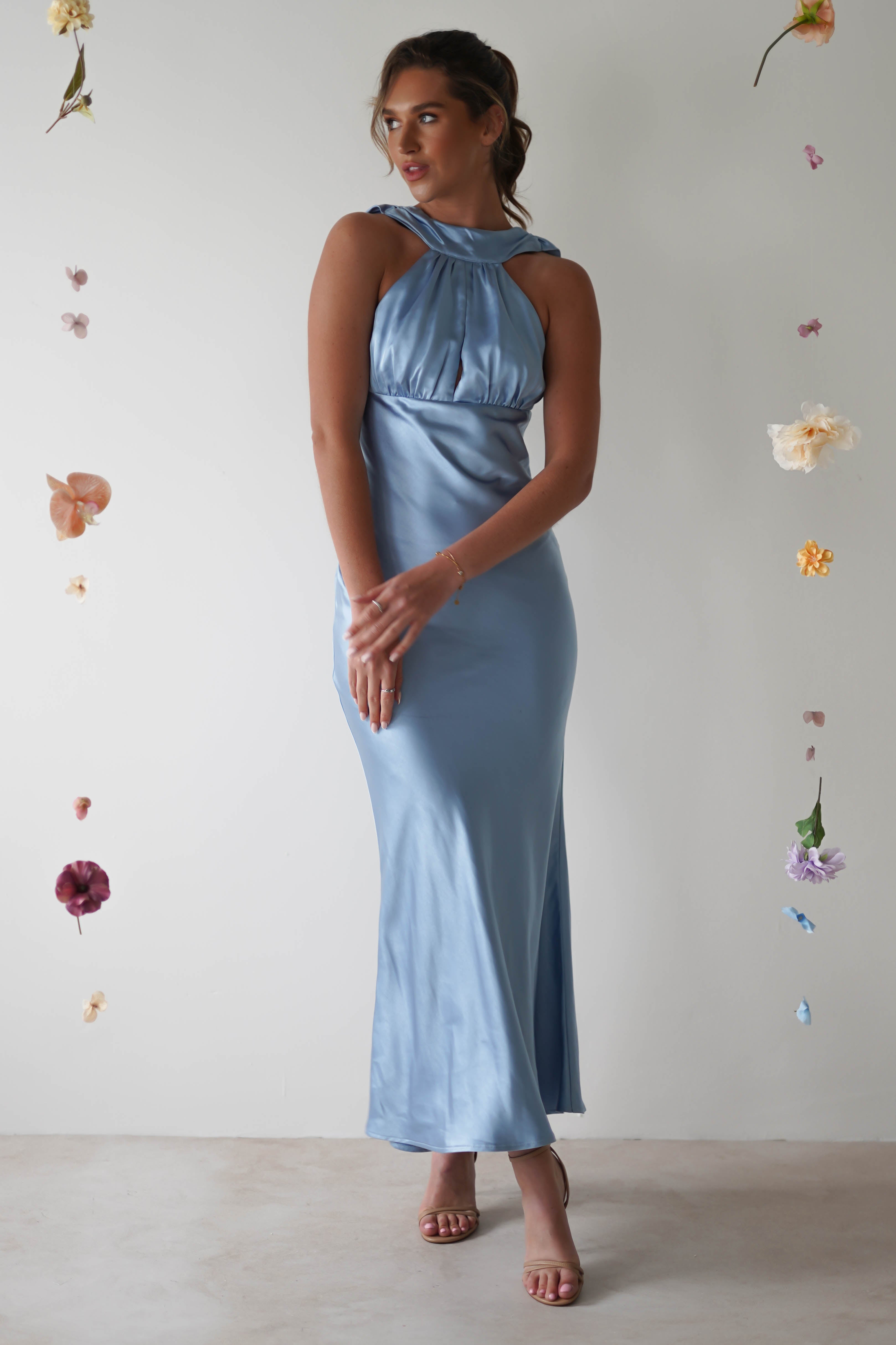 A woman in an elegant light blue satin halterneck maxi dress stands gracefully against a plain background adorned with vertically hanging flowers. She gazes to the side, with her hands lightly clasped in front, revealing an open-back detail that adds a touch of allure.