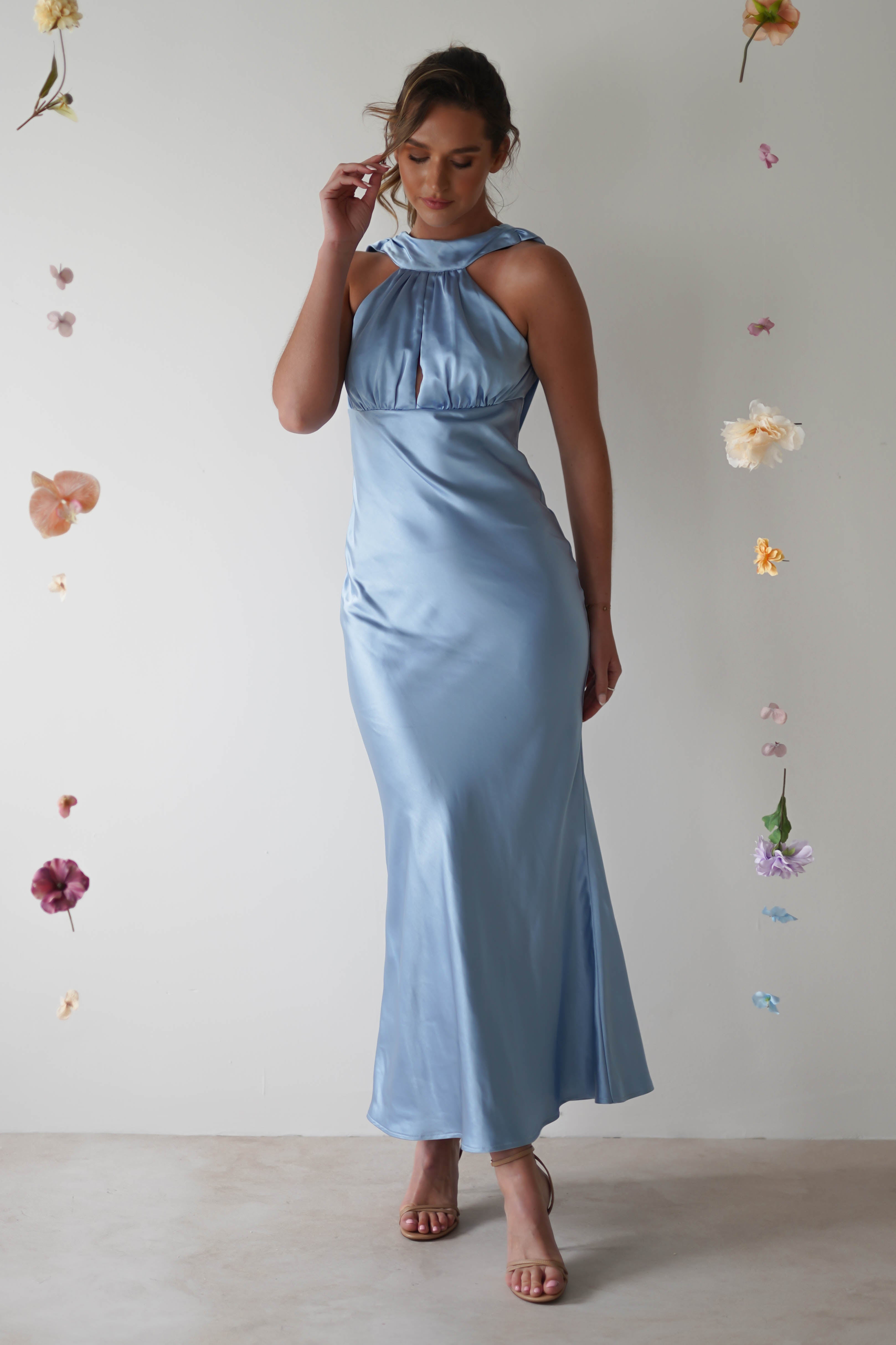 A woman in a sleek, light blue satin gown with an open-back detail stands elegantly. The gown features a unique neckline and a flowy silhouette. She is surrounded by suspended, colorful flowers, creating a whimsical and dreamy atmosphere.