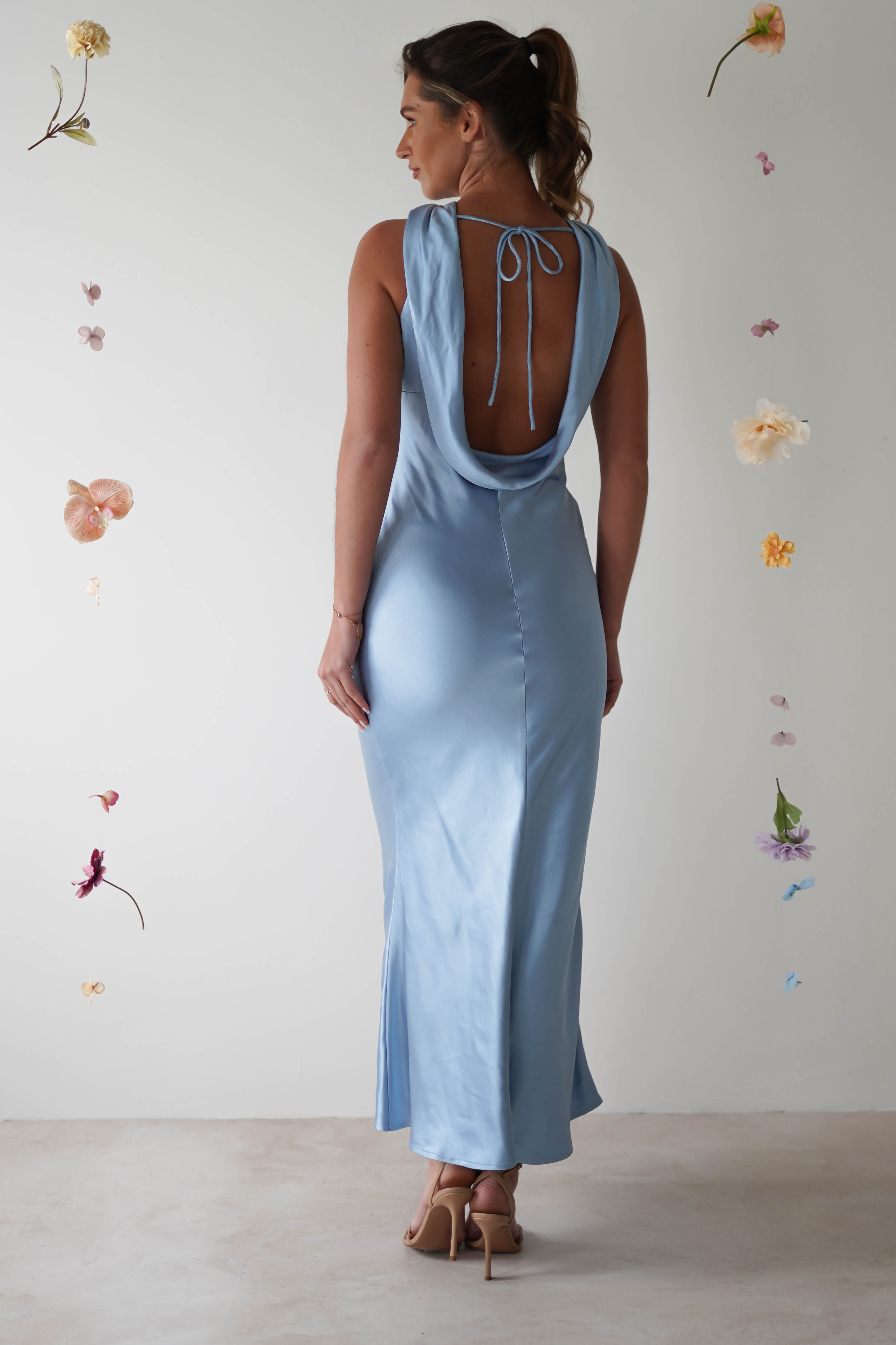 A woman stands facing away, wearing a sleek halterneck maxi dress in light blue satin. The elegant gown features an open-back detail and a tie string at the neck. She is surrounded by delicate, suspended flowers against a plain background.