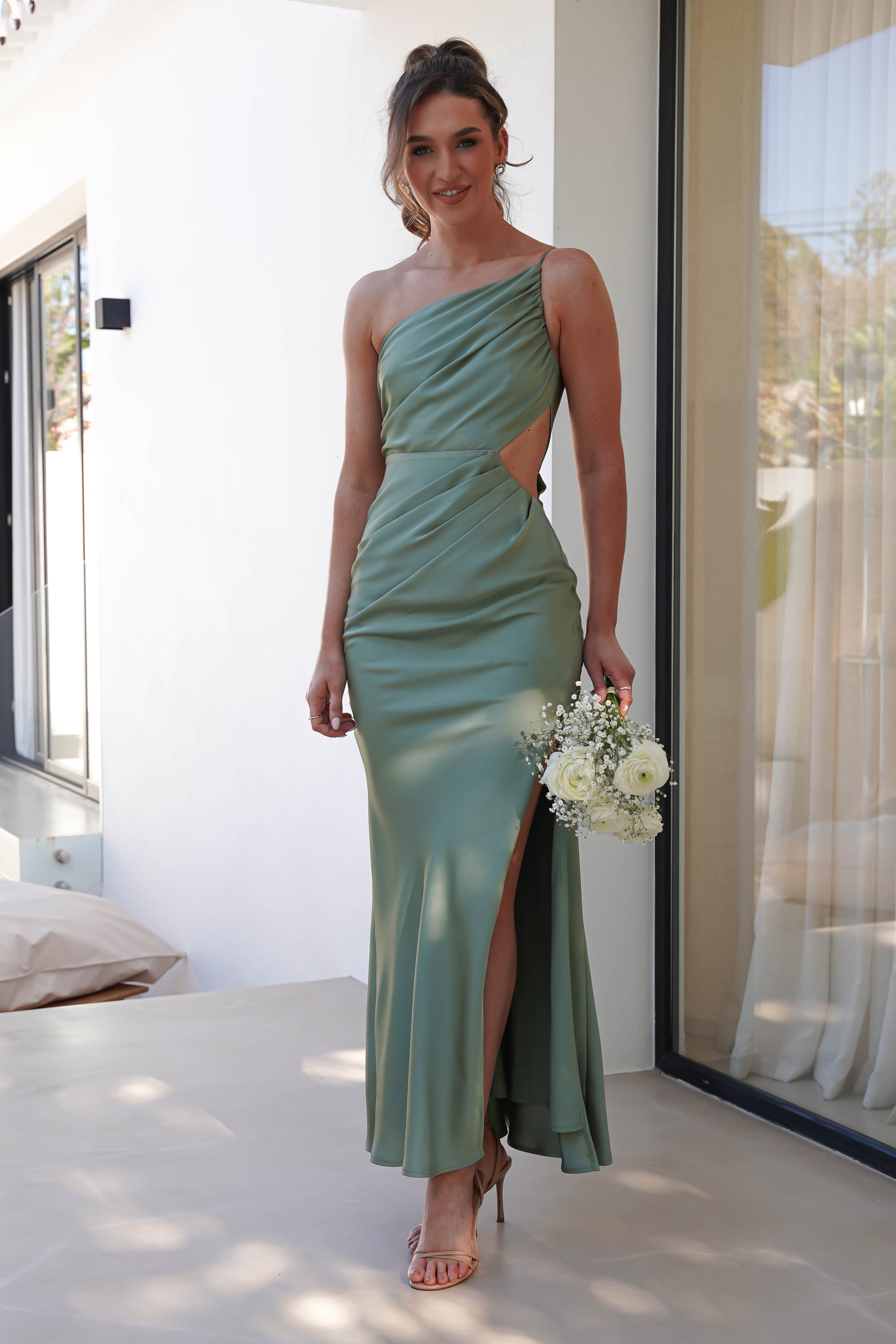 A woman in a sleek, satin maxi dress with side cutouts and a thigh-high slit stands indoors. Shes holding a small bouquet of white flowers and wearing heels. The room, filled with light from the large windows, perfectly complements her elegant wedding guest dress.