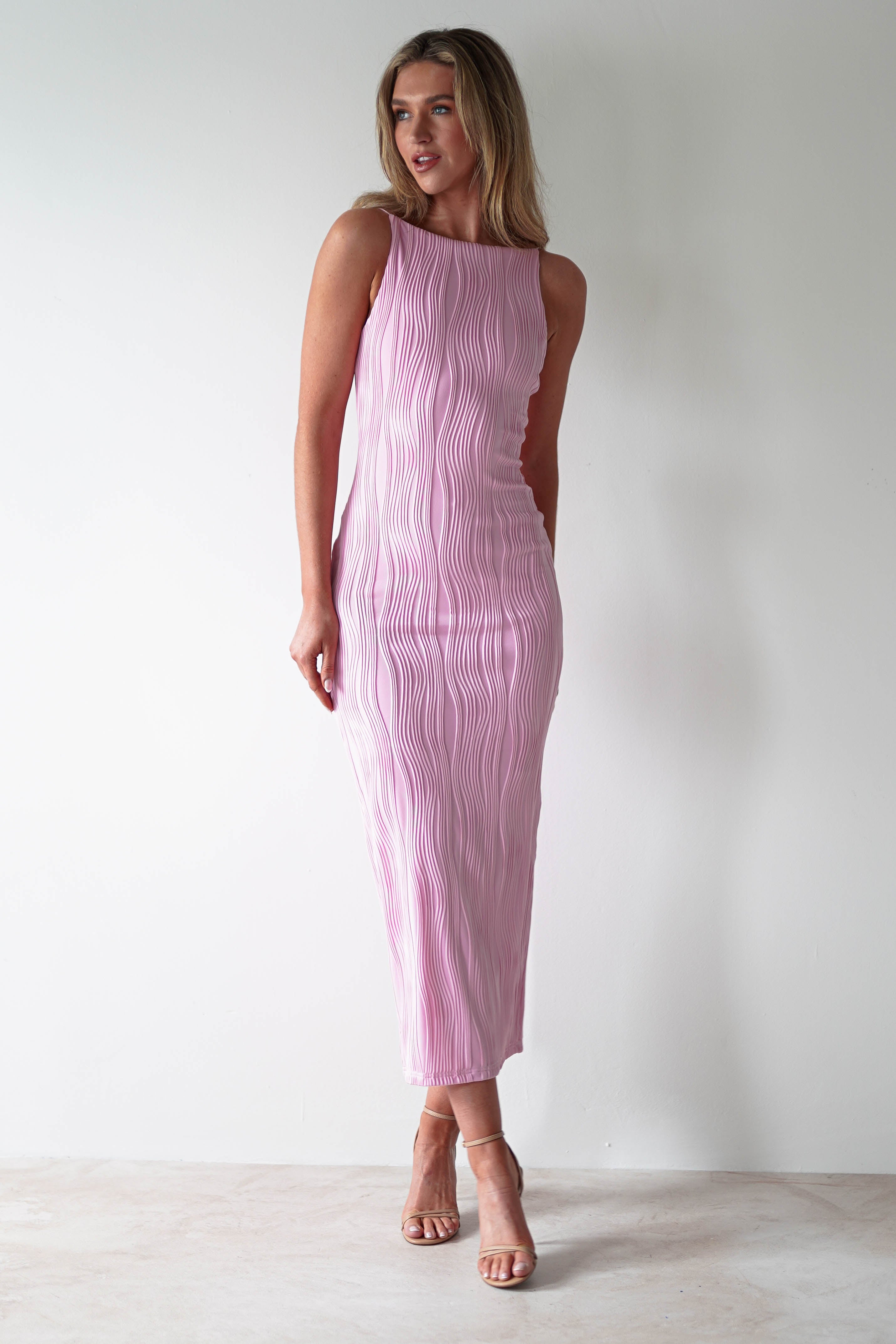 A woman in a stylish textured midaxi dress with wavy vertical patterns stands against a plain white background. Perfect for special occasions, her sleeveless pink ensemble is complemented by nude heels. With long, light brown hair, she gazes to her right with a gentle smile.
