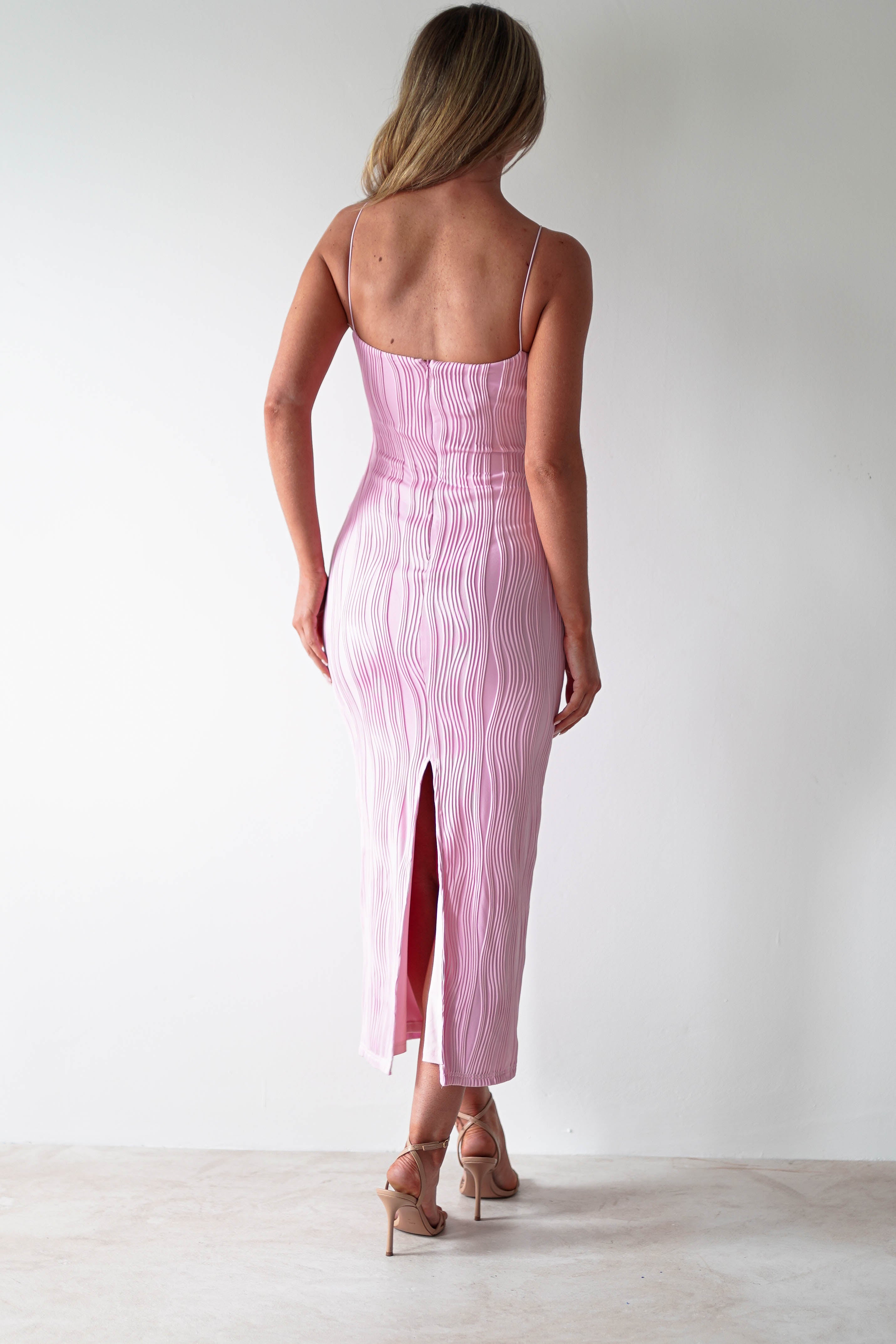 A woman with long hair stands facing a white wall, dressed in a light pink, form-fitting midaxi dress with thin straps and a back slit. The textured fabric complements her beige high heels, making it perfect for special occasions.