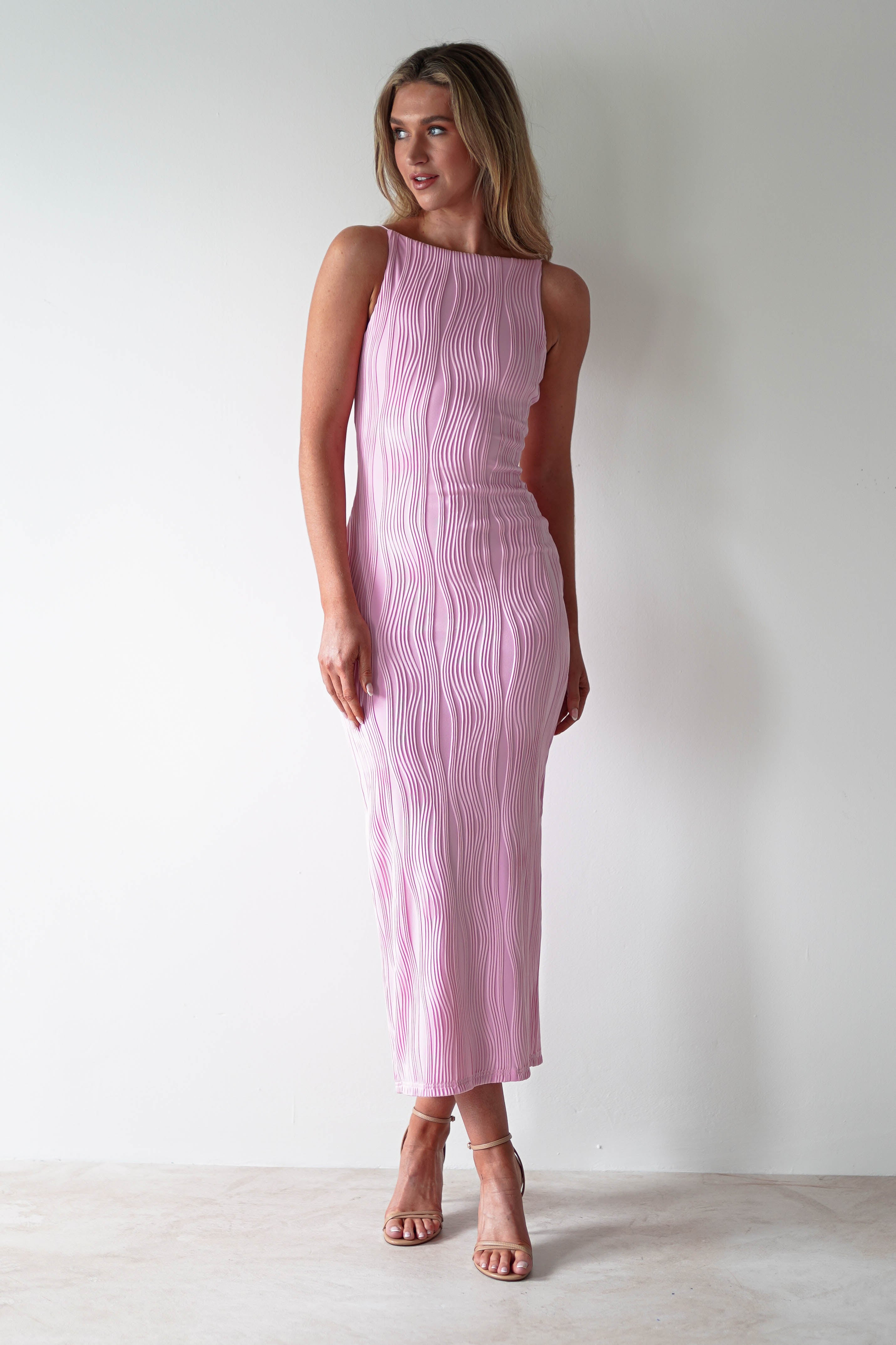 A person in a sleeveless, form-fitting pink midaxi dress stands against a plain white wall. Perfect for special occasions, it features a textured, wave-like pattern. They are wearing beige high heels and have long, light hair.