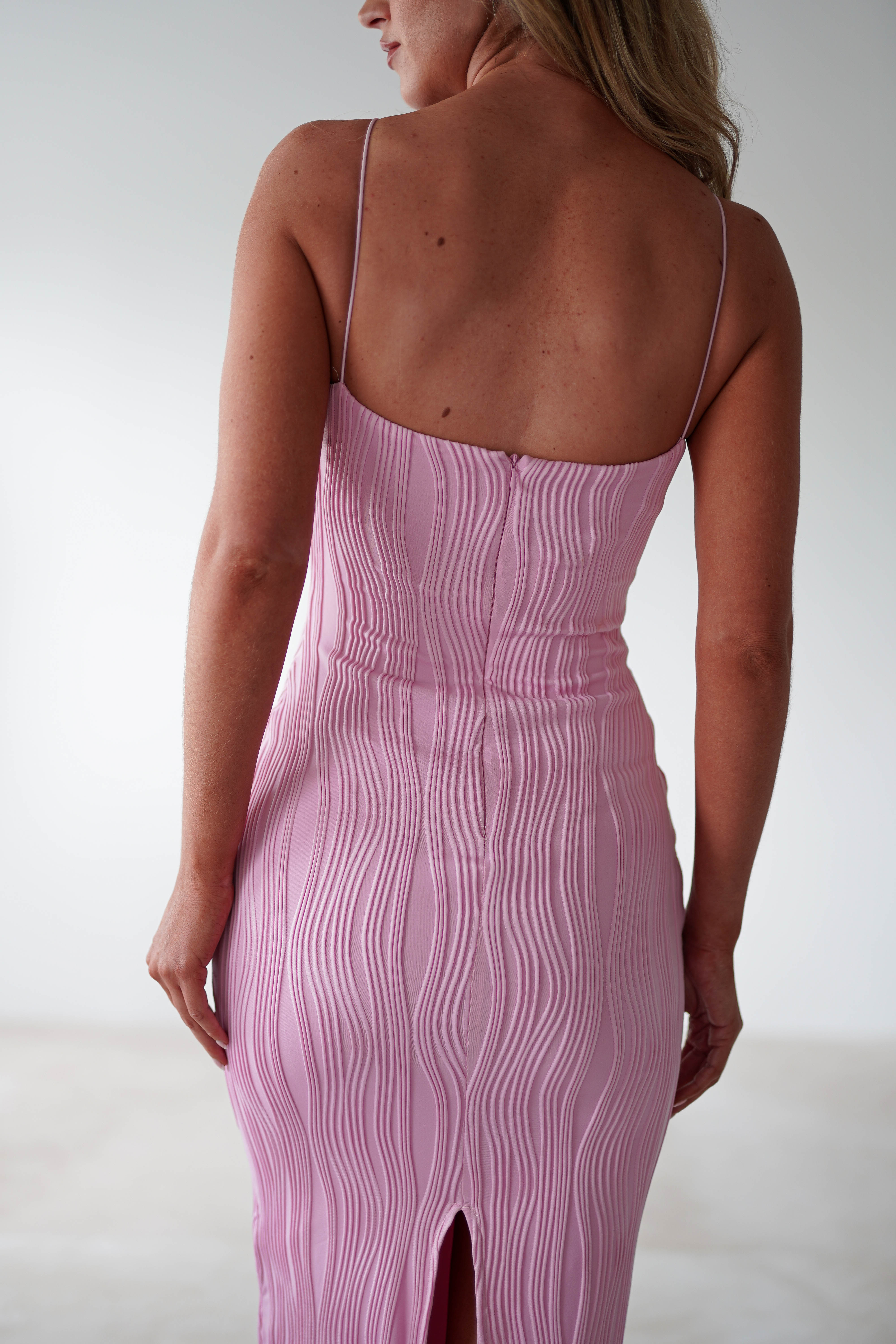 A woman wearing a fitted, light pink midaxi dress with thin straps is standing with her back to the camera. The textured dress features a wavy pattern and a slit at the back. Perfect for special occasions, she stands in a simple, softly lit room.