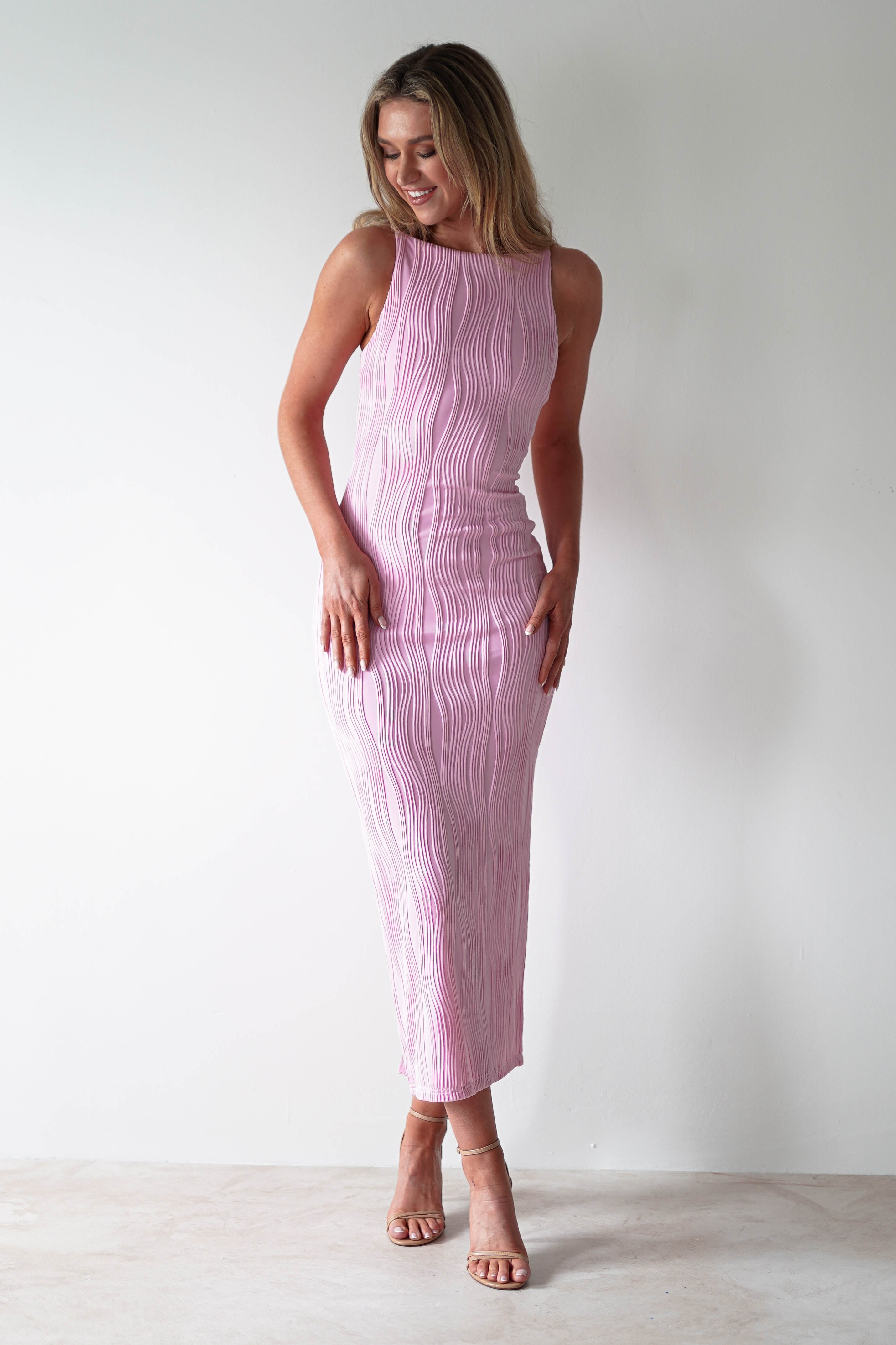 A person wearing a light pink, sleeveless midaxi dress stands against a plain white background. The textured fabric catches the light as they look down and smile, with one hand touching the side of the dress. Neutral heels complete this elegant outfit perfect for special occasions.