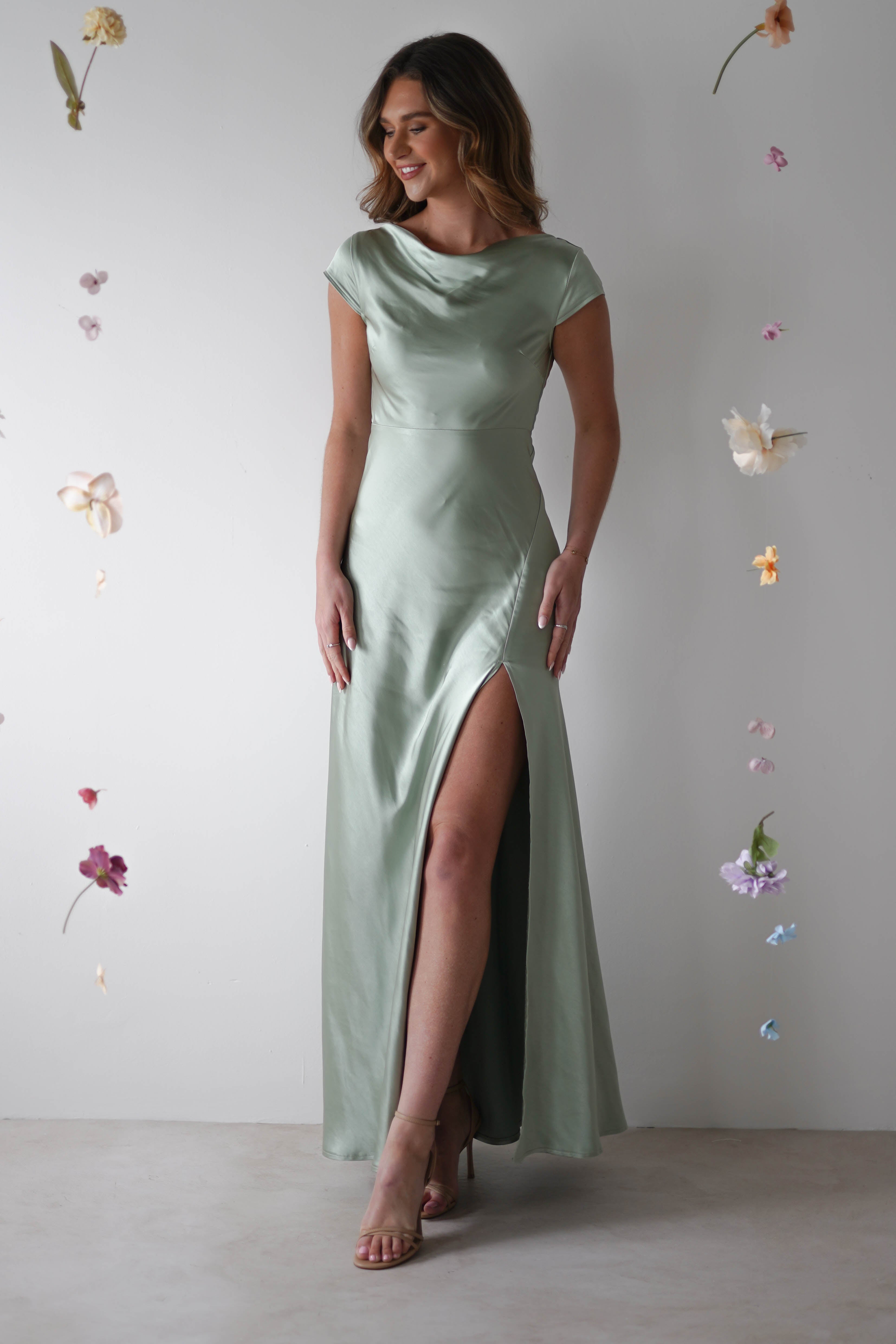 A woman wearing a light green satin maxi slip dress with a thigh-high slit stands against a white background, surrounded by hanging pastel flowers—perfect for bridesmaids or as a chic wedding guest look.