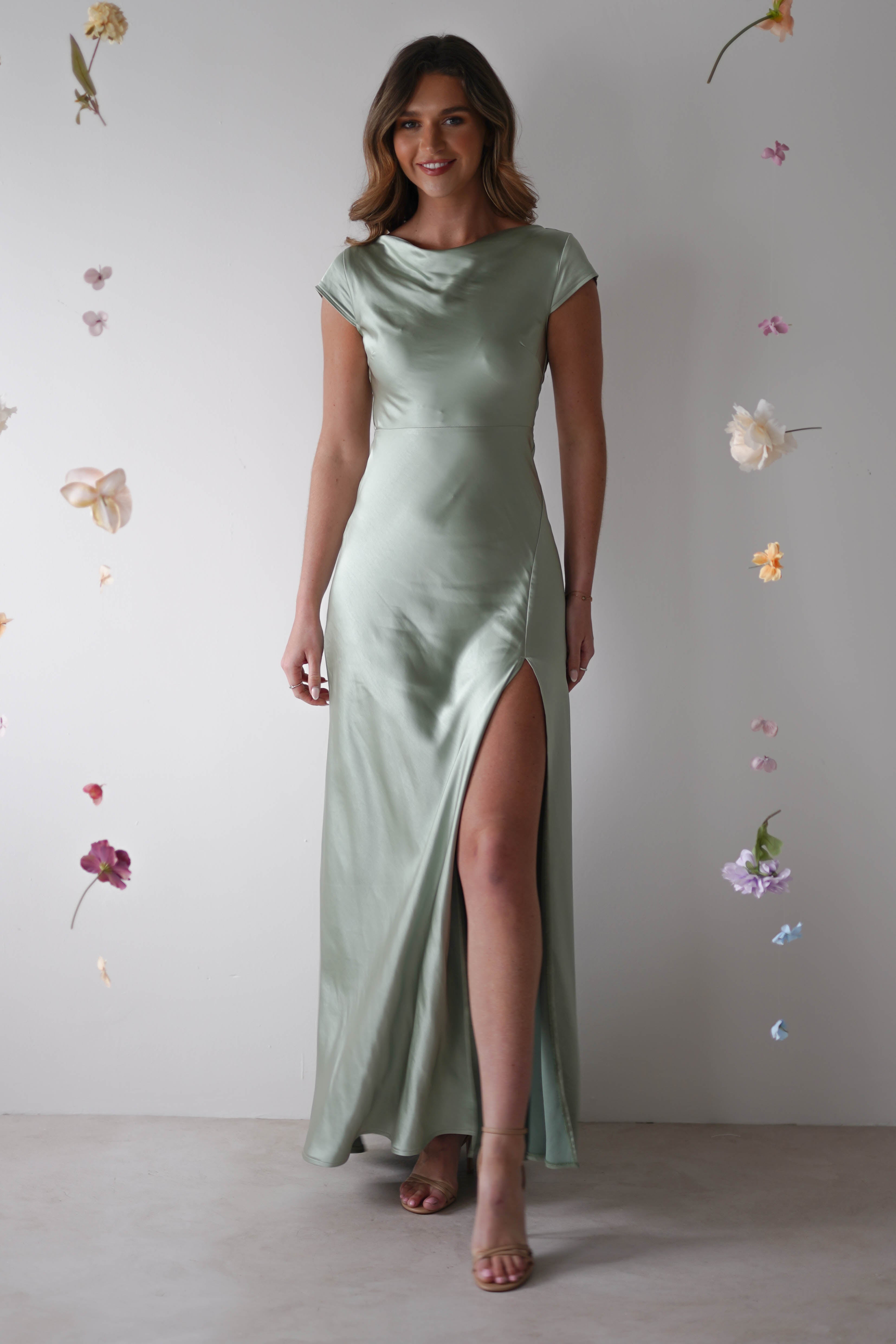 A woman wearing a light green satin maxi slip dress with a thigh-high slit stands against a white background decorated with floating pastel flowers—a perfect look for bridesmaids or any stylish wedding guest.