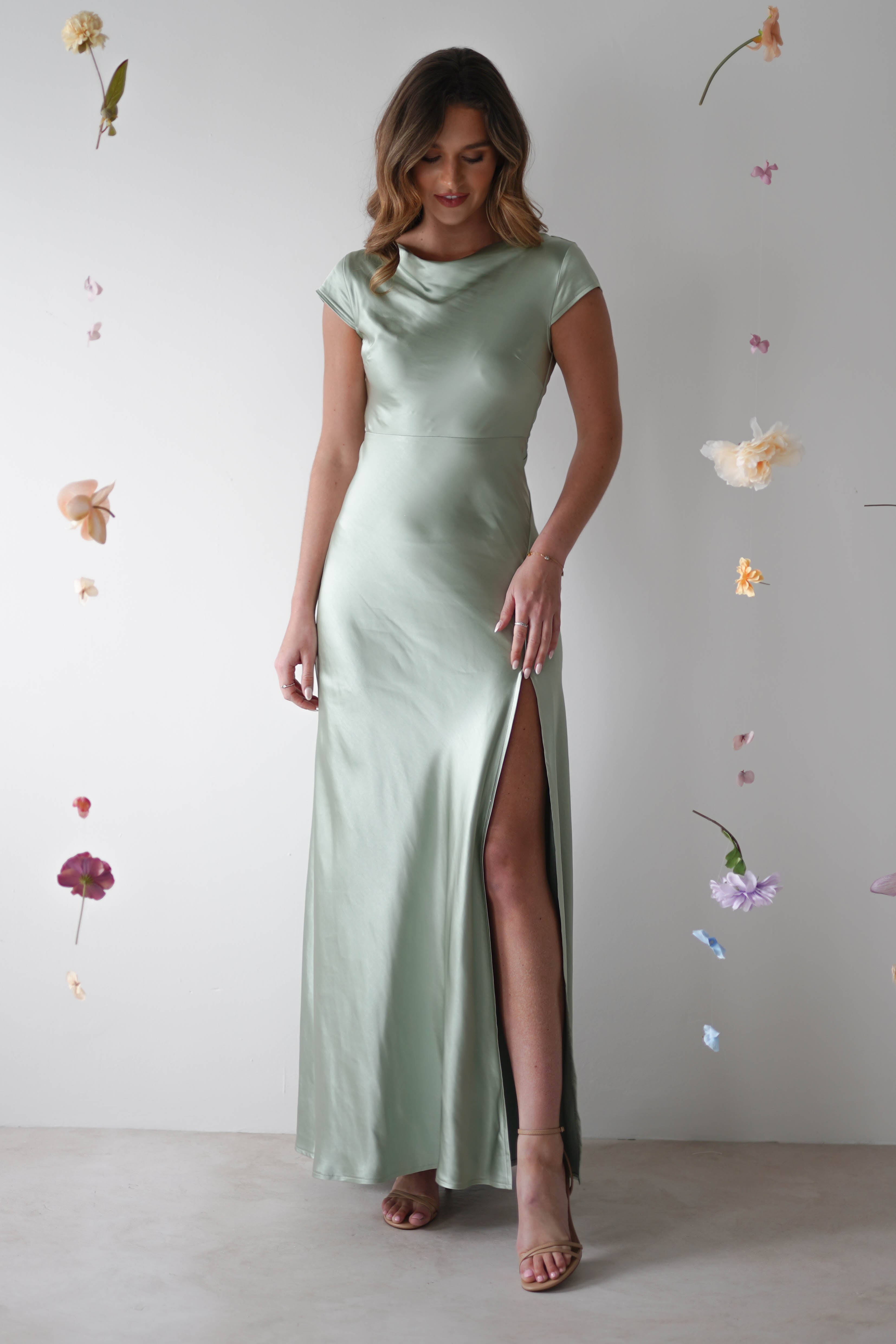 A woman wearing a light green, short-sleeved, floor-length satin maxi slip dress with a thigh-high slit stands against a white background, surrounded by hanging flowers—perfect for wedding guest or bridesmaids attire.