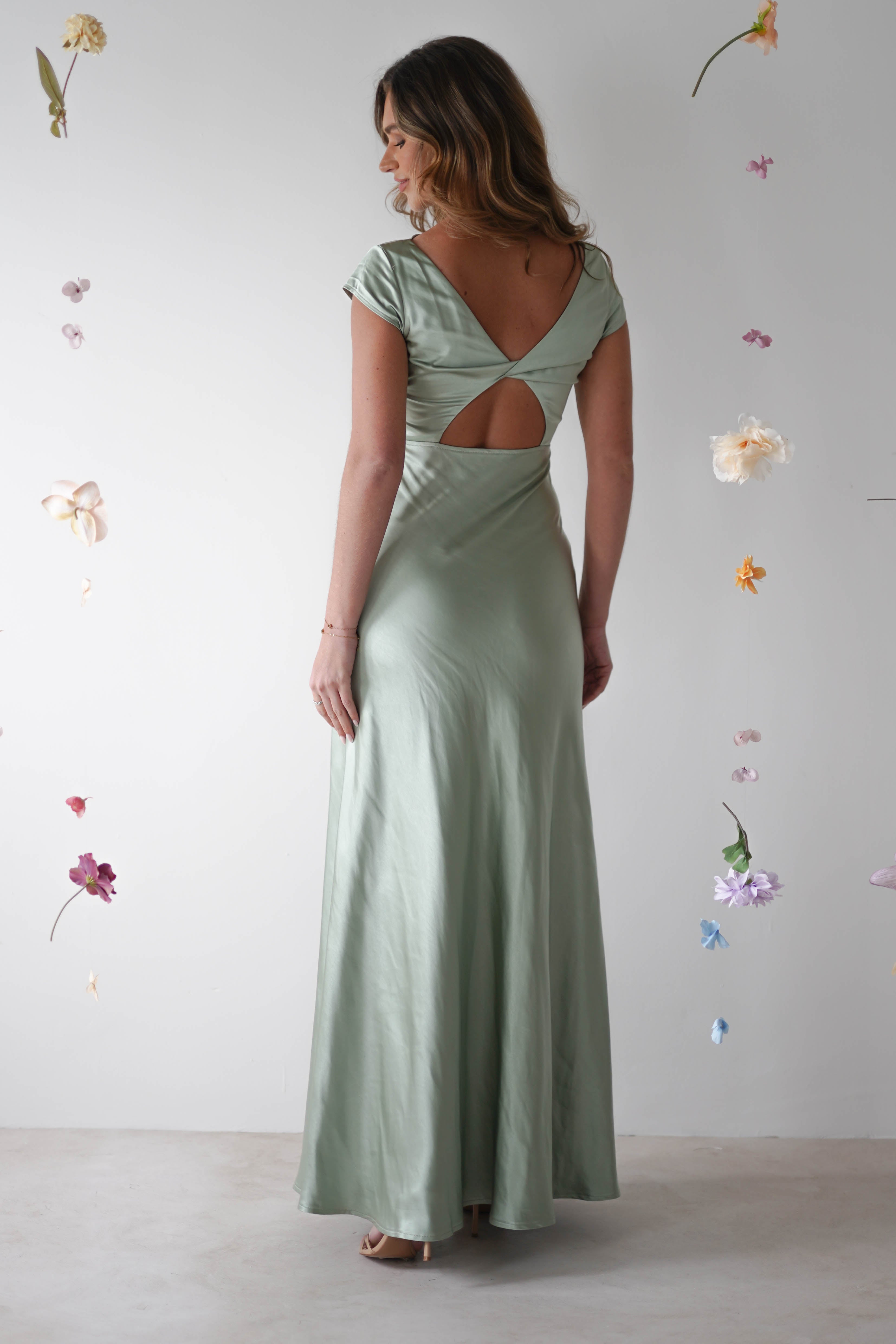 A woman in a light green maxi slip dress with a cutout back stands facing away, surrounded by floating flowers—a perfect look for bridesmaids or a stylish wedding guest against a plain white background.