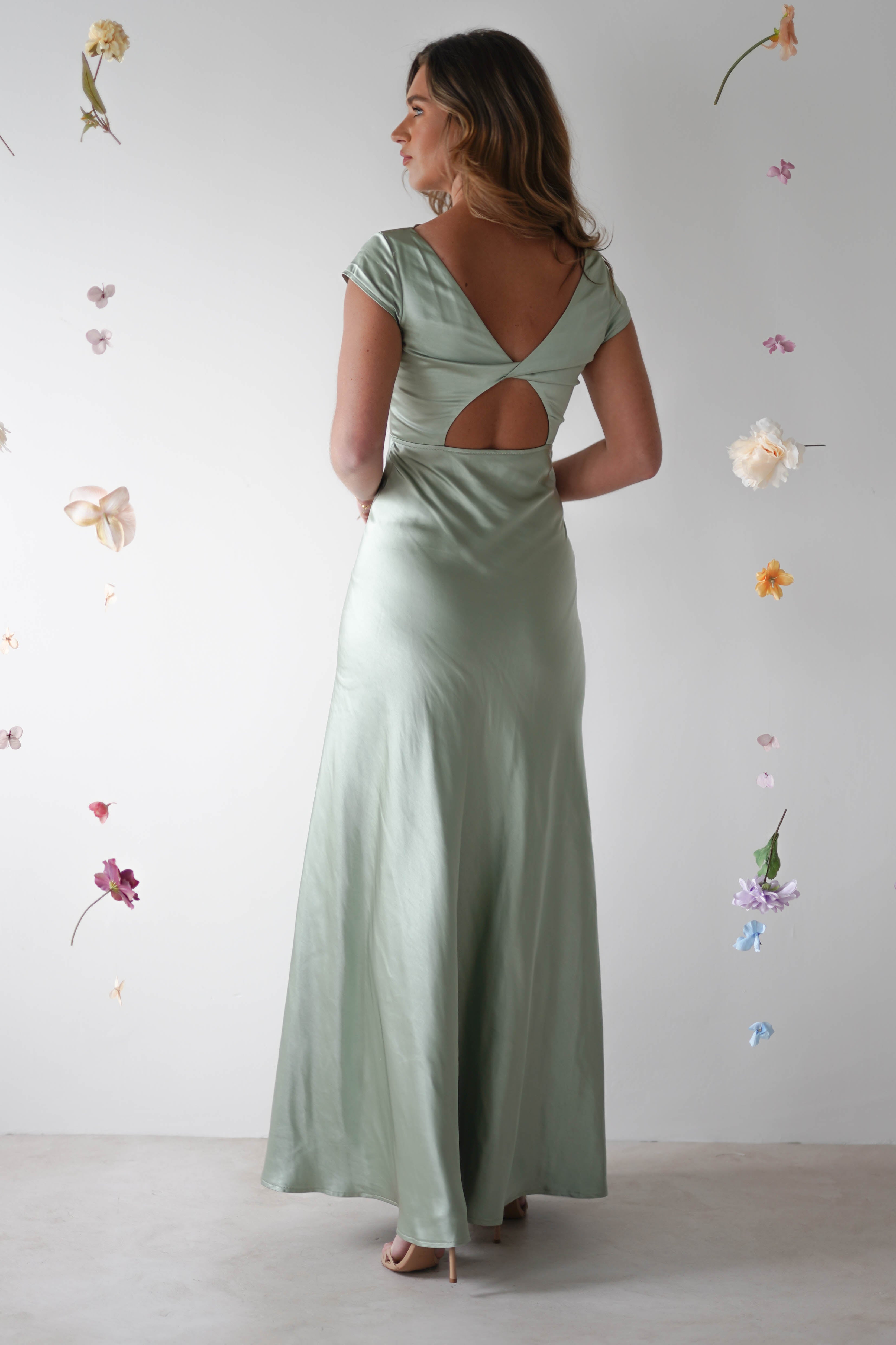 A woman in a light green satin maxi slip dress with a V-shaped open back and cap sleeves stands facing away. Perfect for bridesmaids or as a wedding guest look, she is surrounded by floating pastel flowers on a plain white background.