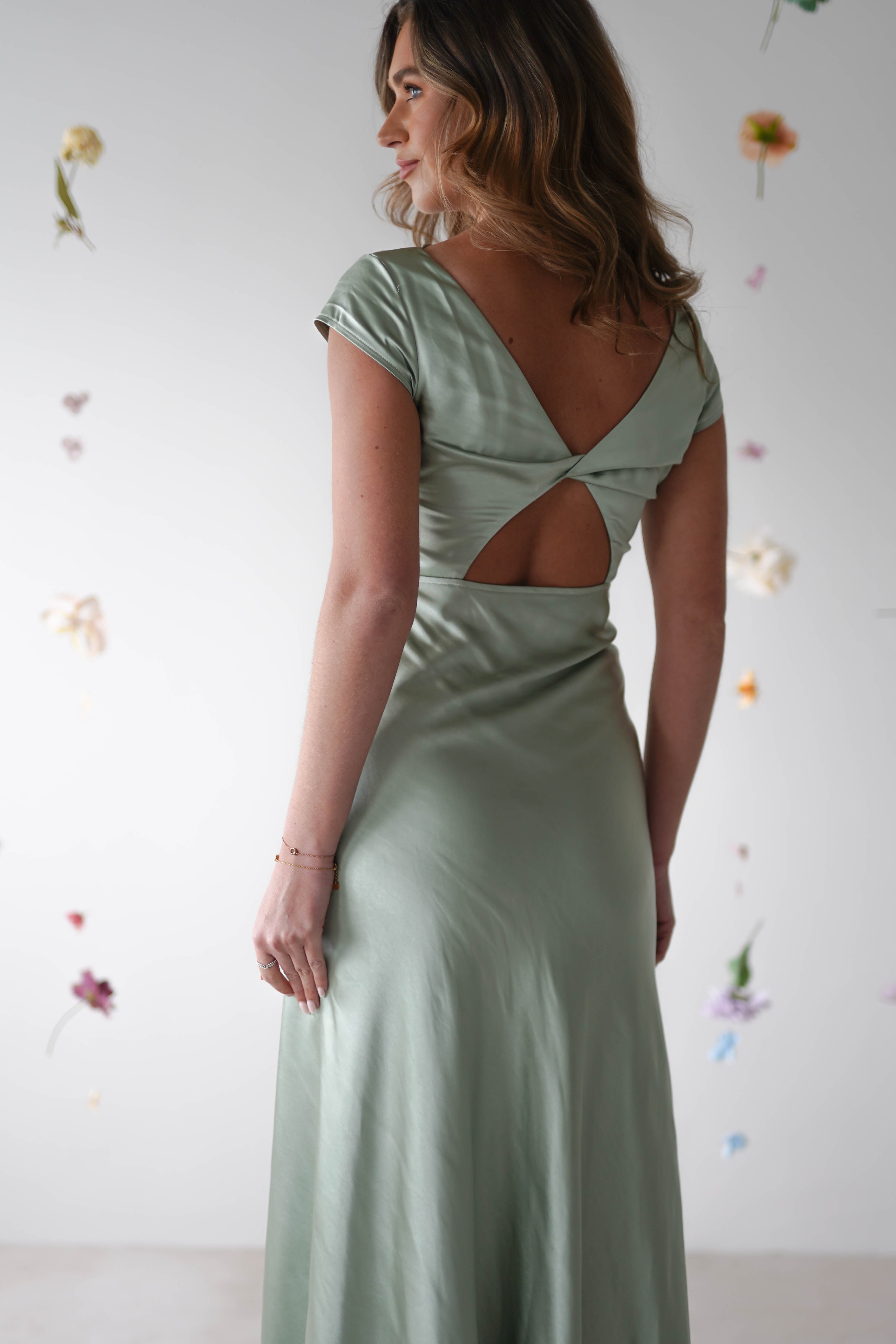A woman wearing a light green maxi slip dress with a V-back and cutout detail stands facing away, perfect for bridesmaids or a wedding guest, with soft light and scattered flowers hanging in the background.