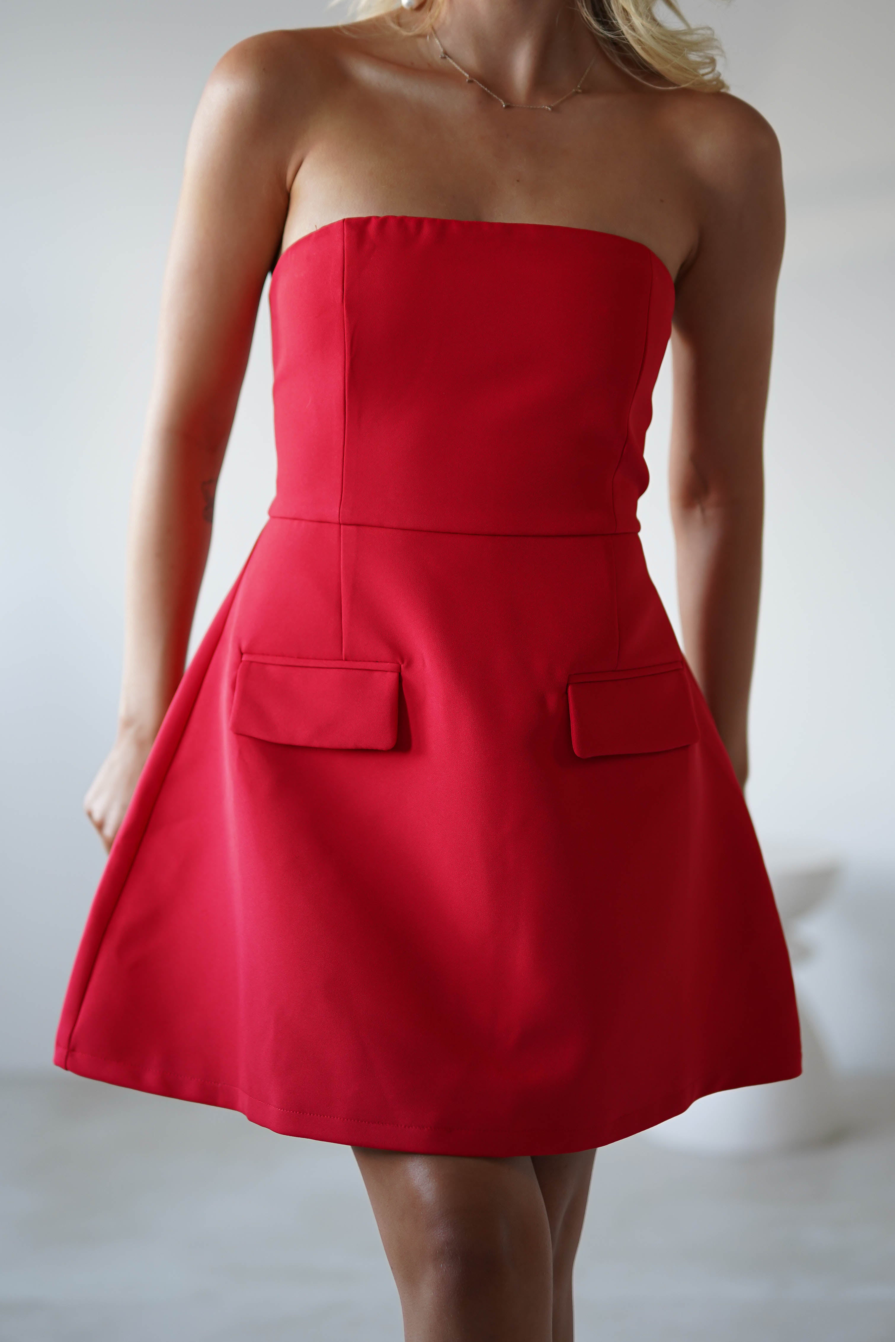 A woman in a strapless mini red dress flares out slightly, showcasing two rectangular pockets at the front. Perfect for wedding guests or graduations, the background remains neutral and elegantly blurred.