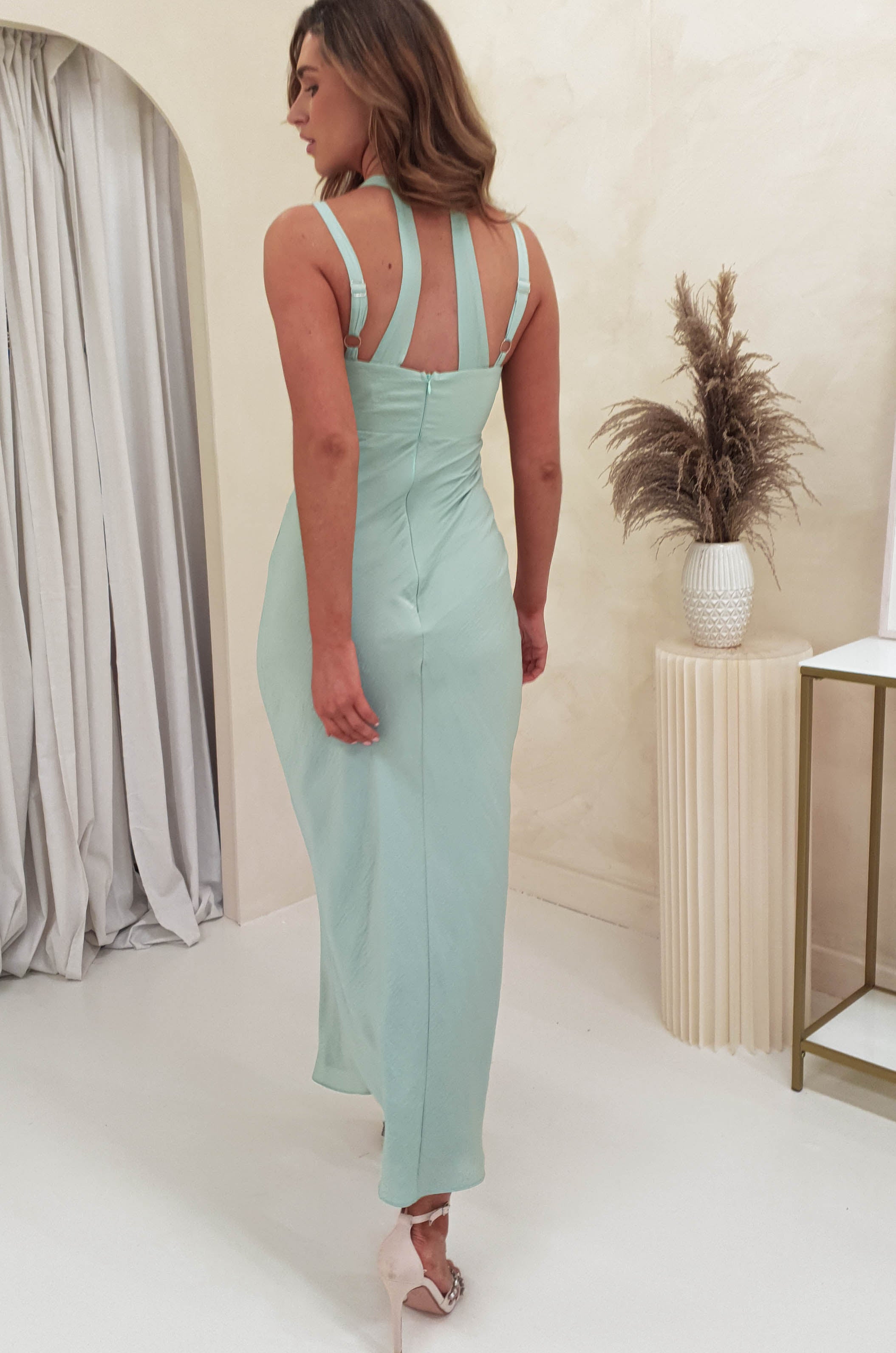 A woman in a long, pale blue dress with adjustable straps poses in a room with light-colored curtains and a vase holding dried grasses. She is wearing high heels on the white floor, beside a small side table.