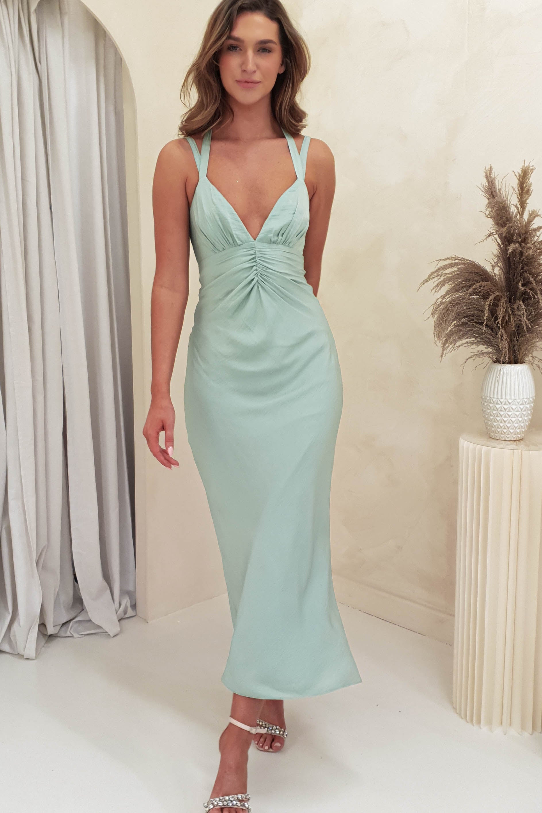 A woman in a light green, ruched body dress with adjustable straps poses gracefully in a room with light-colored walls and curtains. Standing near a round beige table adorned with pampas grass, she complements her look with strappy silver heels.