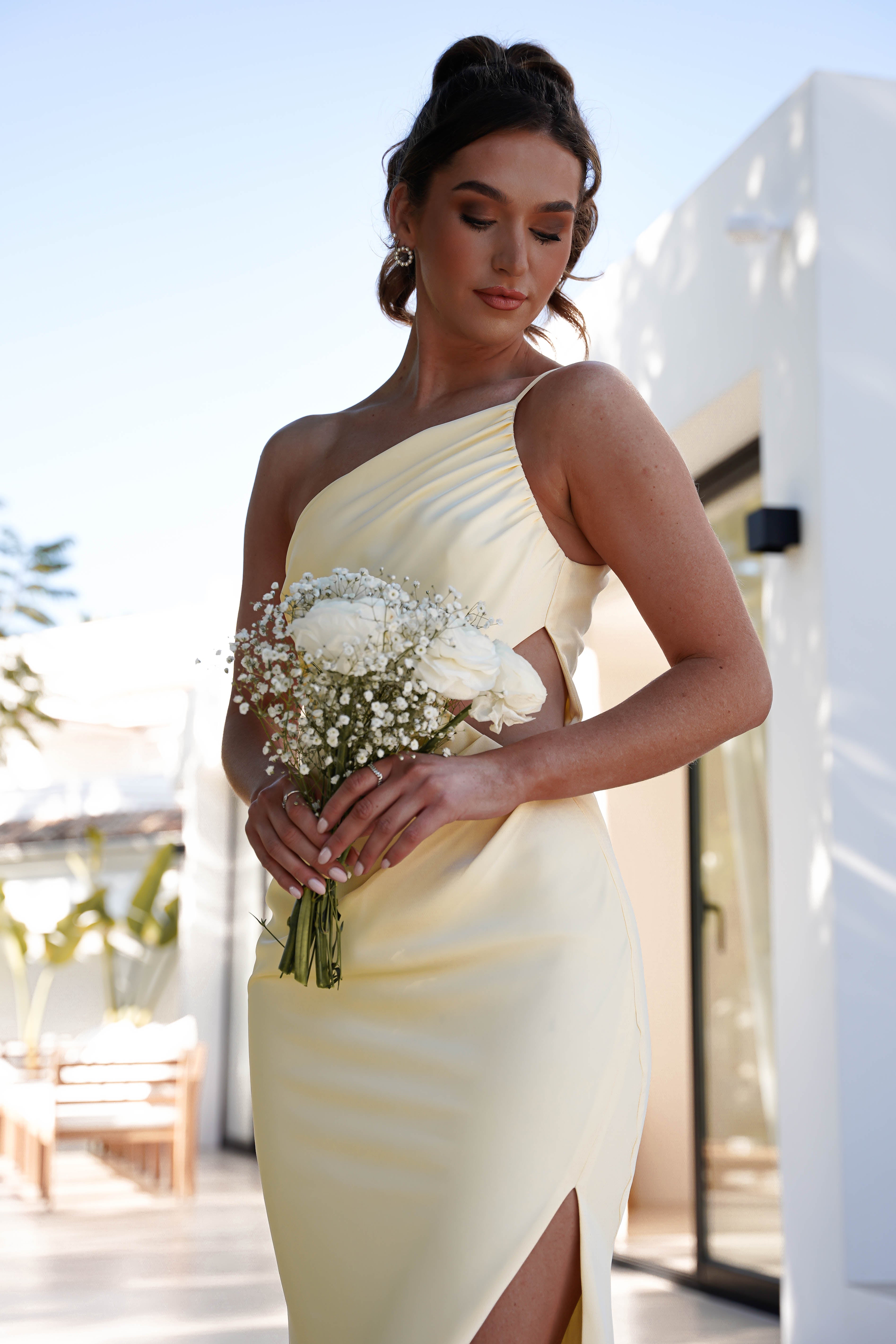 A woman in a yellow one-shoulder satin maxi holds flowers, radiating elegance and charm.