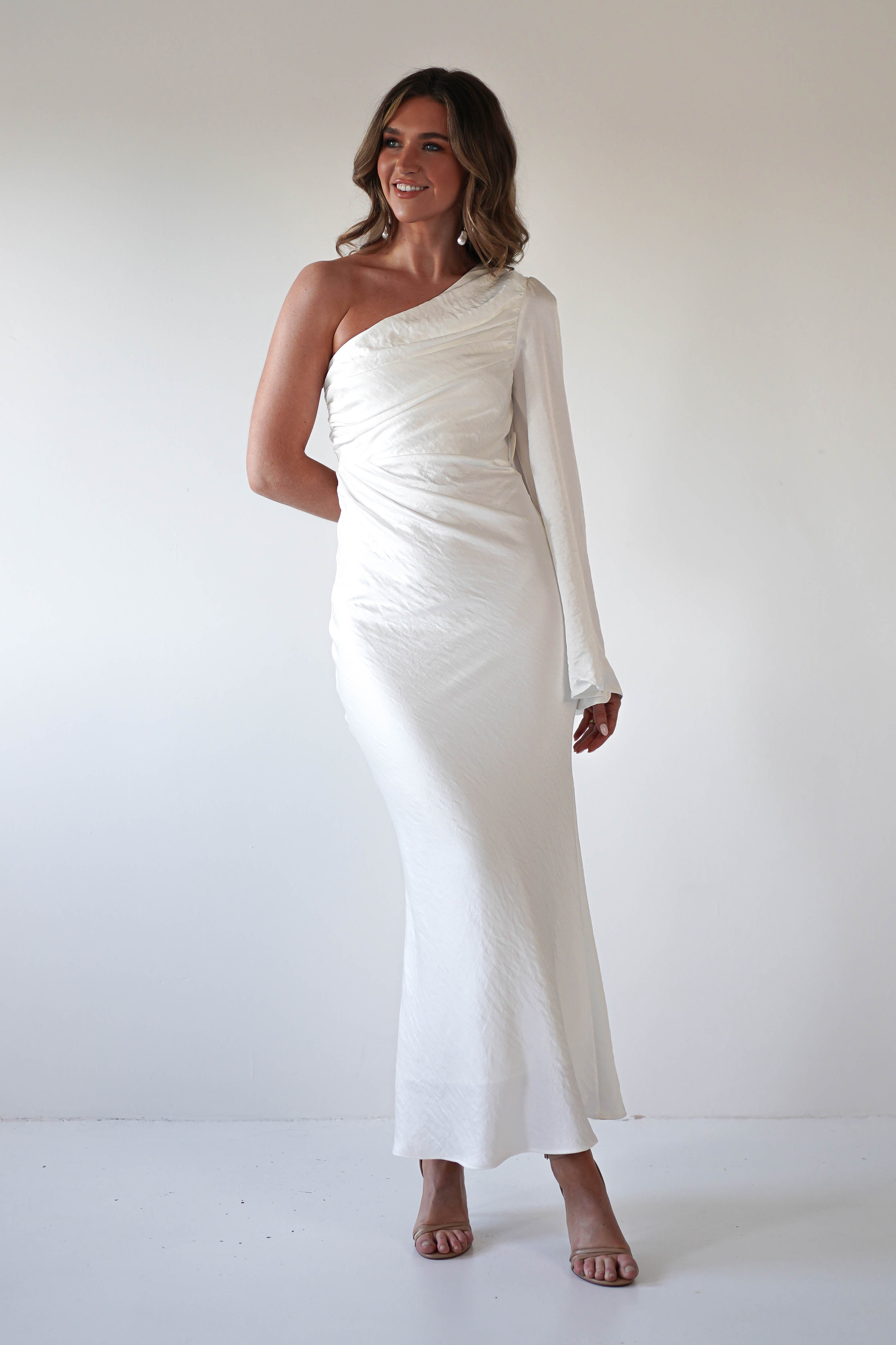 A woman stands against a plain white background wearing an elegant ensemble—a long, white formal dress with an asymmetrical neckline and single long sleeve. She has wavy hair and is smiling, with one hand by her side and the other behind her back.