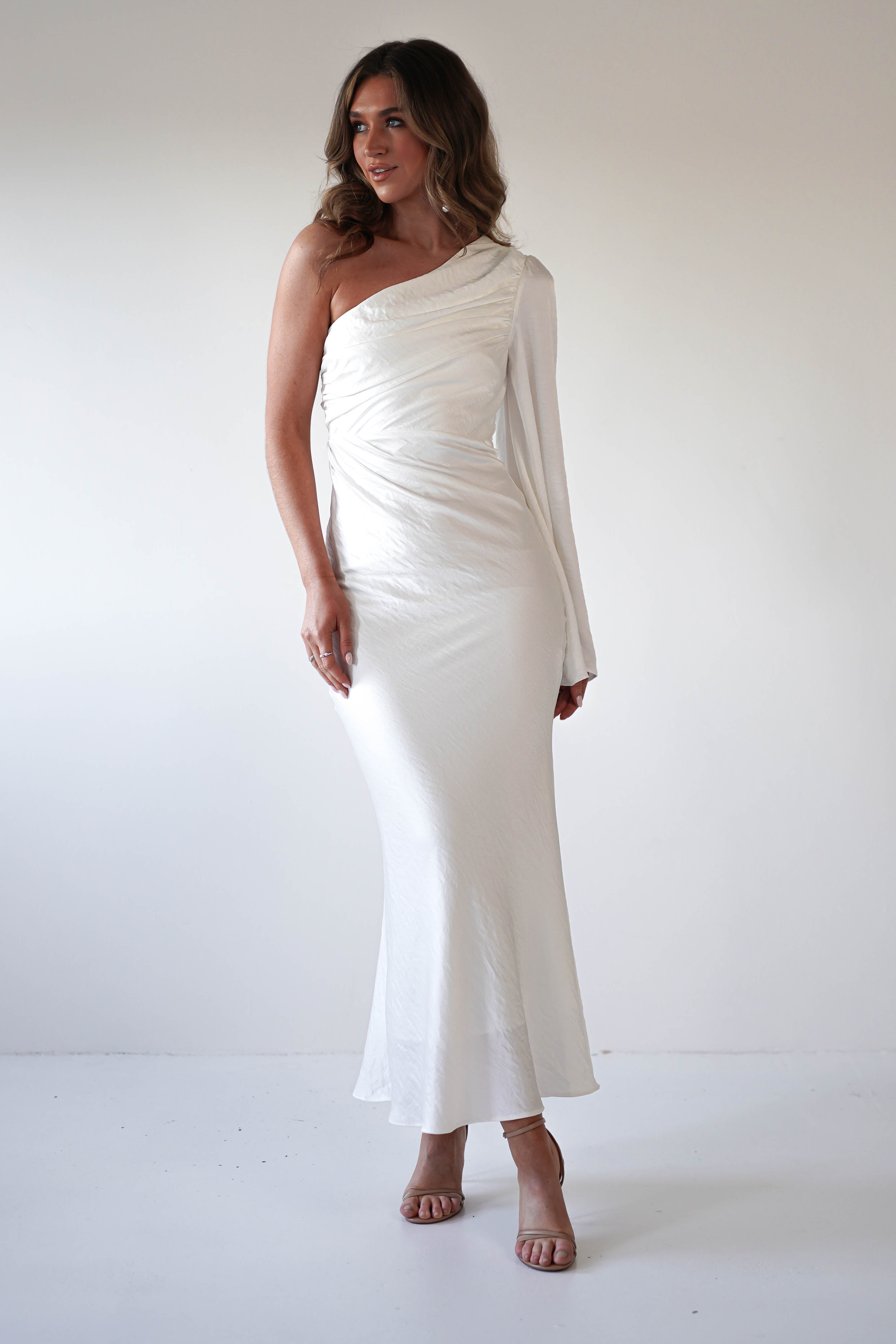 A woman stands against a plain white background wearing a formal dress with an asymmetrical neckline—one long sleeve and one bare shoulder. Her elegant ensemble is completed with long, wavy hair and nude high-heeled sandals.