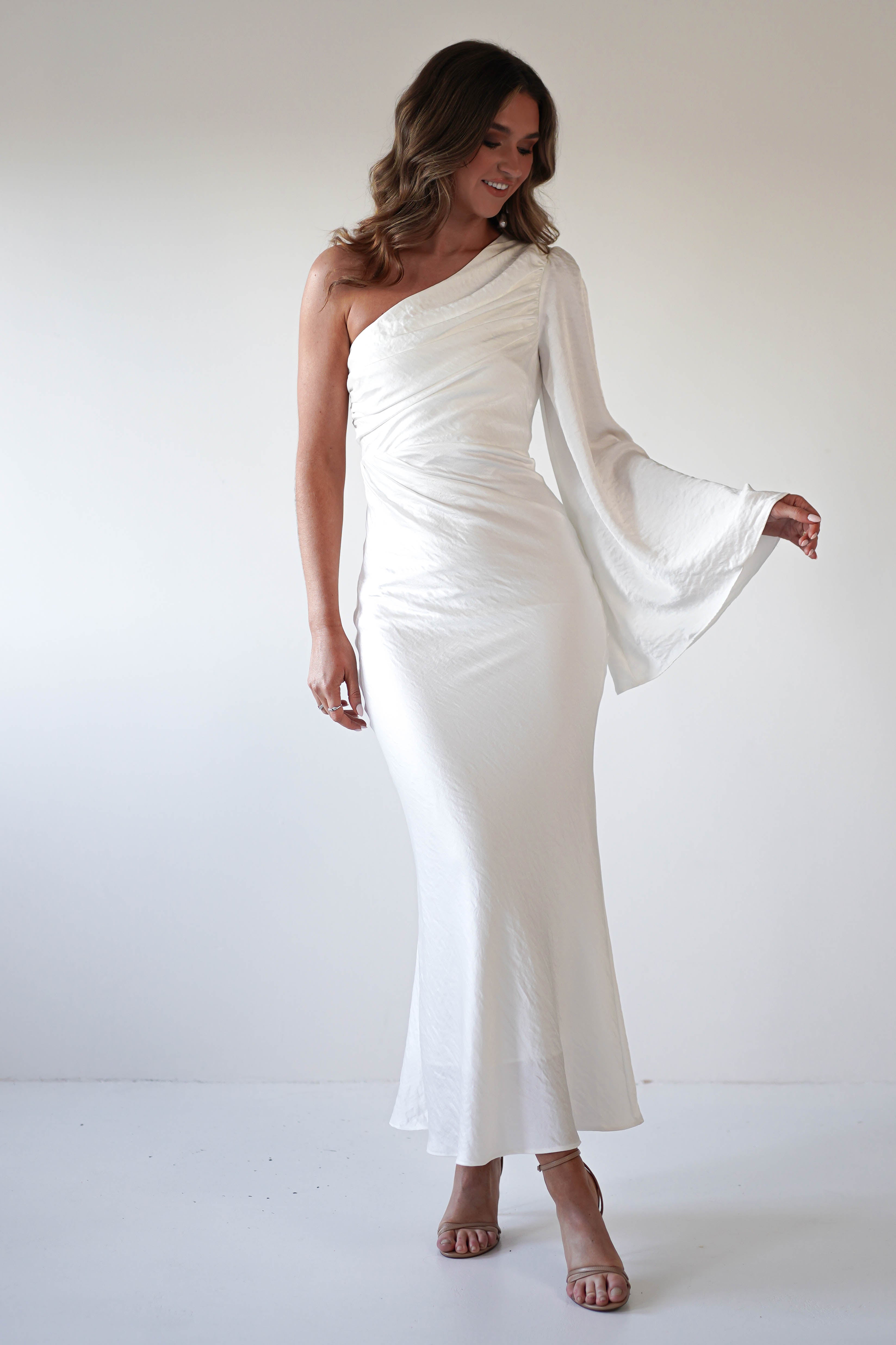 A woman wearing a long, white formal dress with an asymmetrical neckline and a flared sleeve stands against a plain background, looking down and smiling slightly.