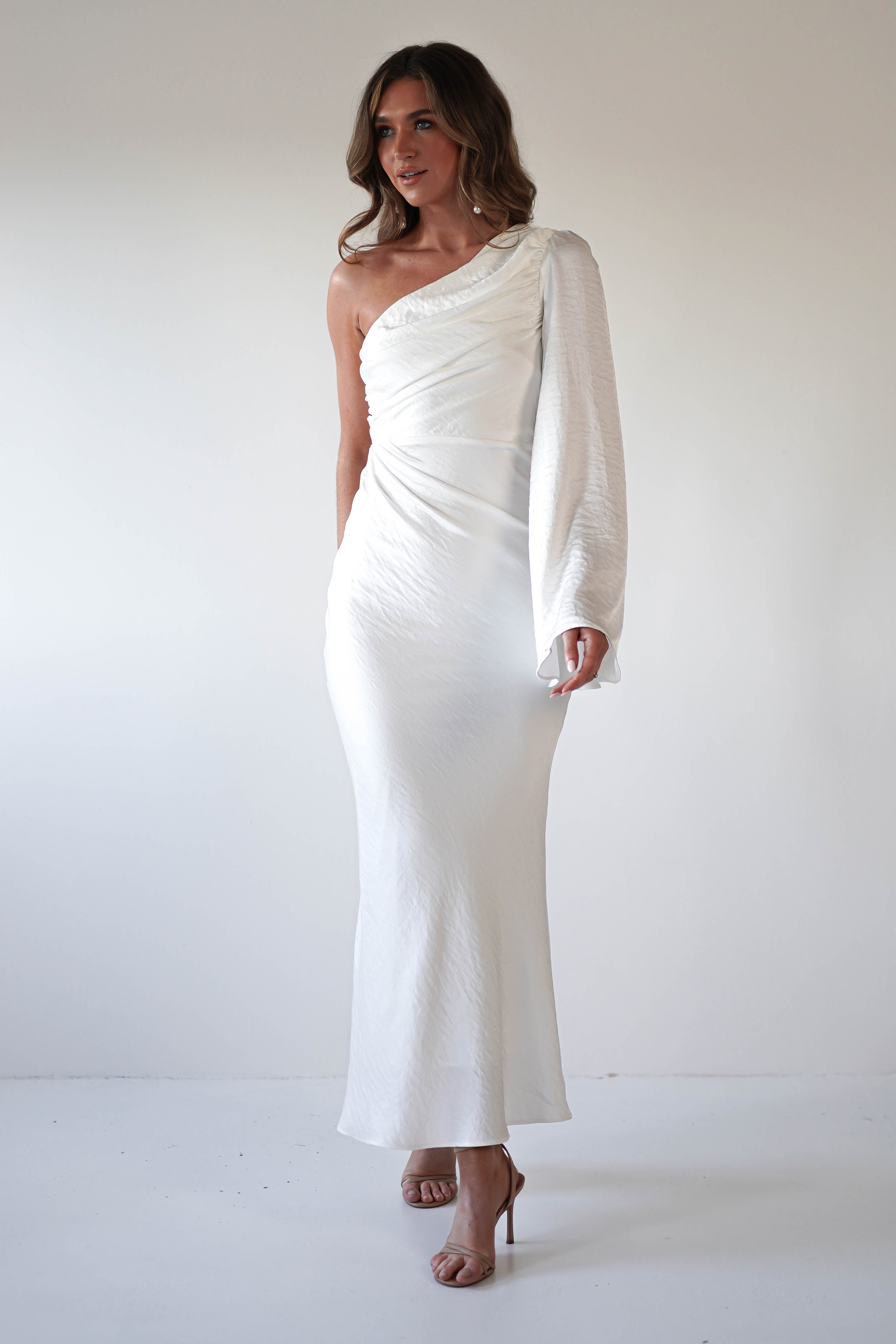 A woman stands against a plain white background, wearing an elegant ensemble: a long, white formal dress with an asymmetrical neckline, draped sleeve, and fitted silhouette. She has wavy brown hair and wears nude high heels.