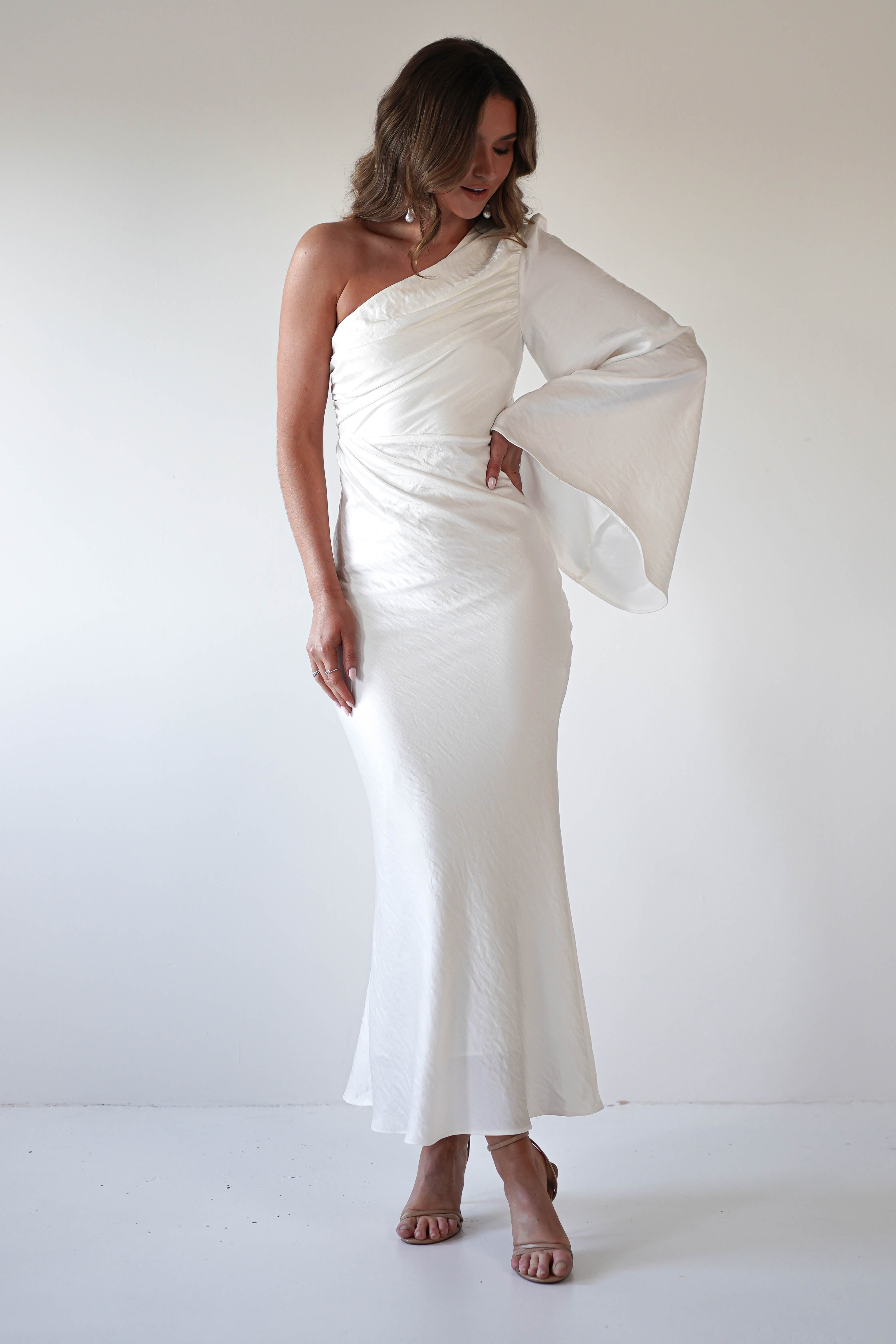 A woman in a long, white formal dress with an asymmetrical neckline and flared sleeve poses against a plain white background, looking down with one hand on her hip and wearing open-toe heels.