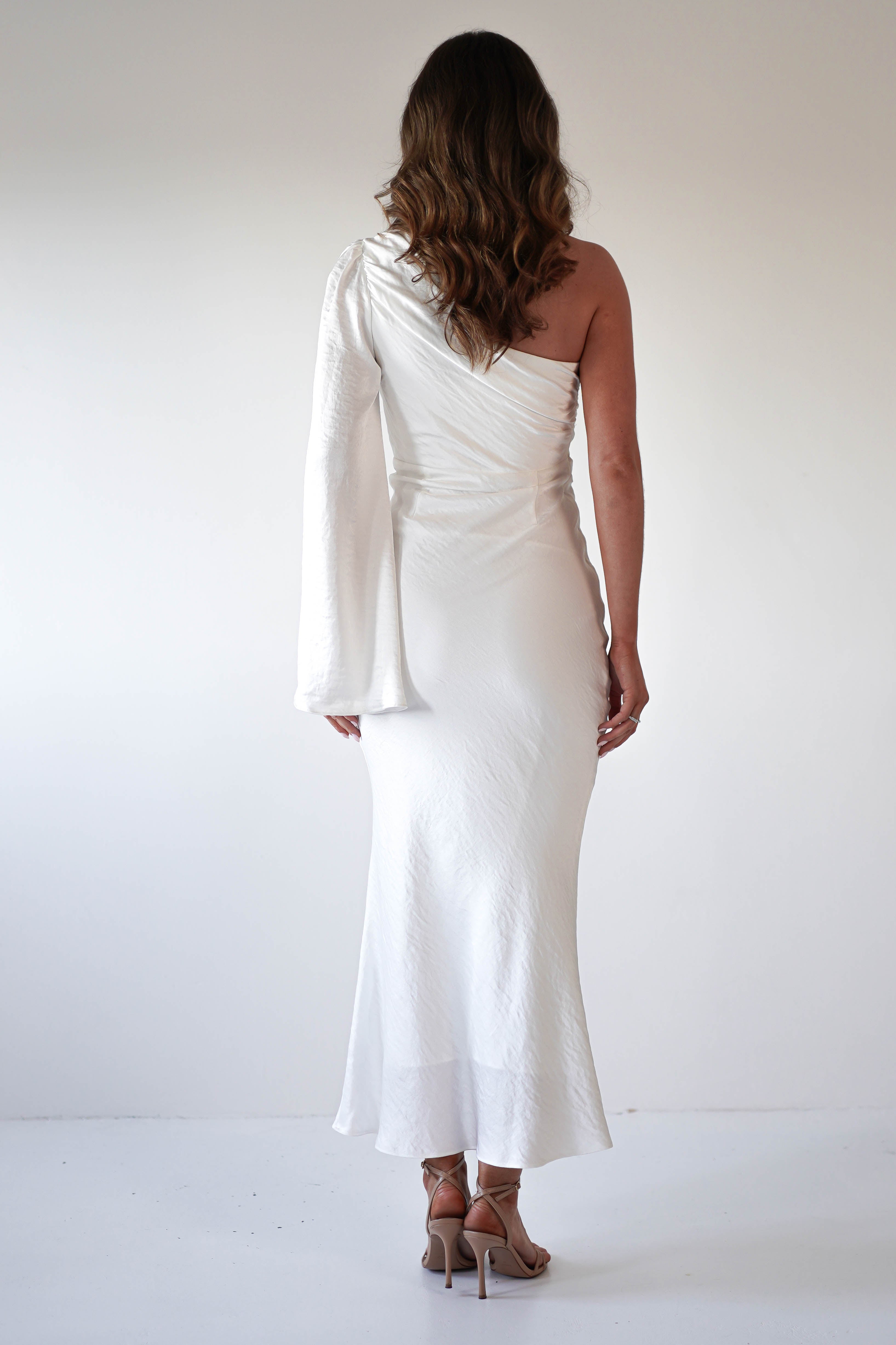 A woman with wavy brown hair stands facing away, wearing an elegant ensemble—a white formal dress with an asymmetrical neckline and long sleeve, paired with beige high-heeled sandals against a plain white background.