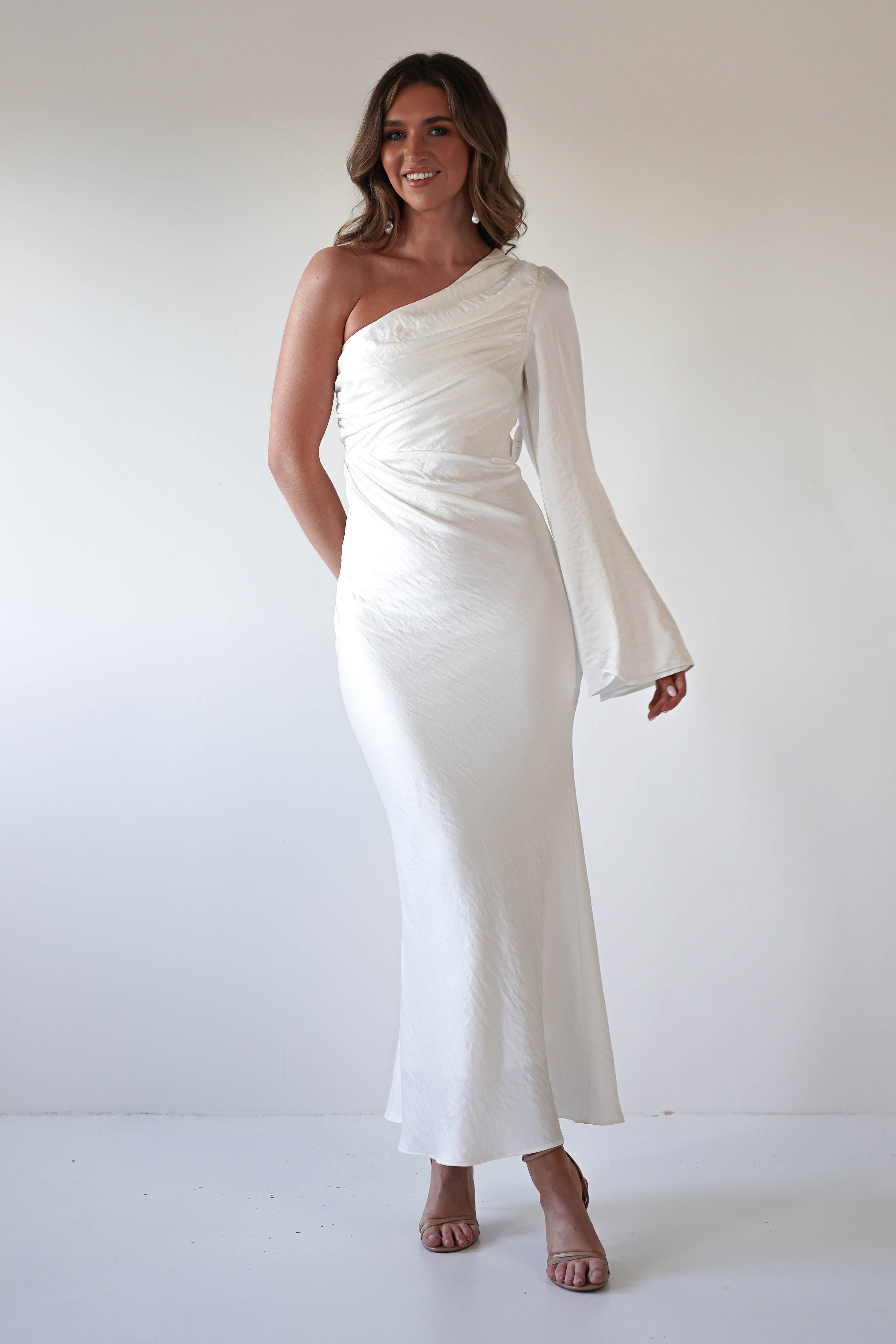 A woman wearing a formal dress with an asymmetrical neckline and flared sleeve poses against a plain white background, smiling with her hand at her side and her hair in loose waves.