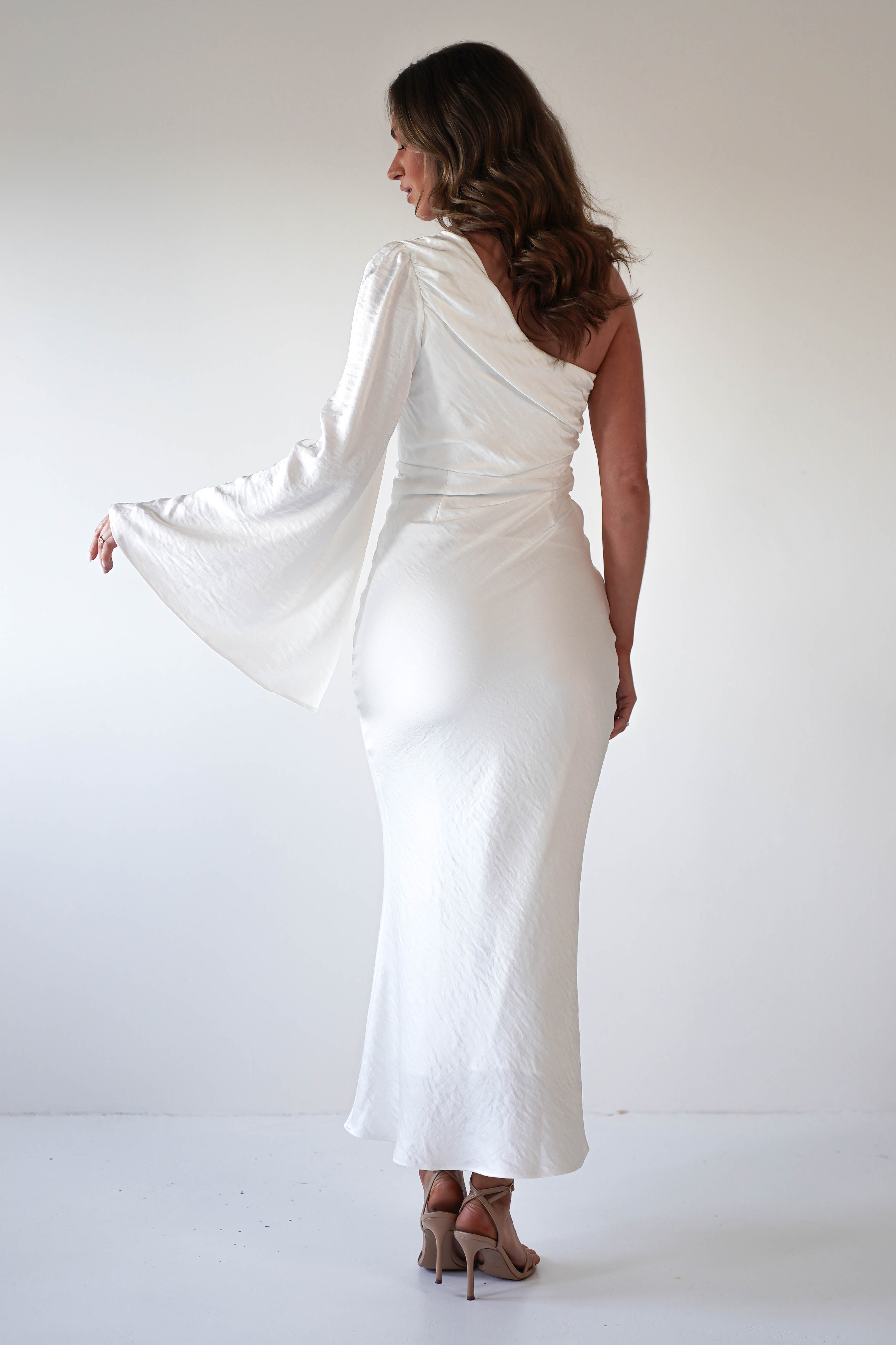 Woman in a white, formal dress with an asymmetrical neckline and flared sleeve stands facing away, looking to the left. She has wavy brown hair and wears high-heeled sandals against a plain background.