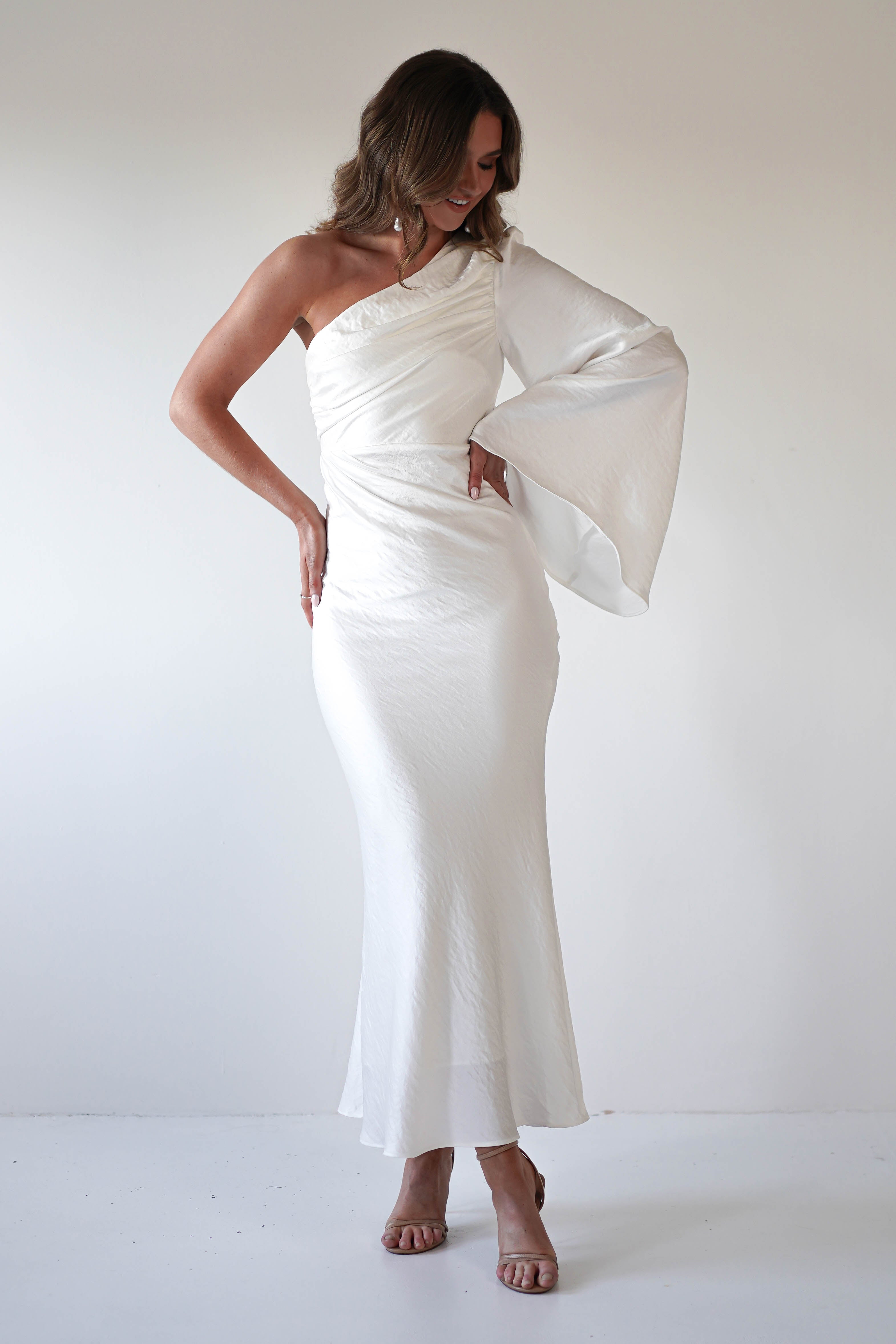 A woman stands against a plain white background wearing an elegant ensemble—a long, white, one-shoulder formal dress with a dramatic flared sleeve. She looks down and smiles, with one hand resting on her hip and the other on her thigh.