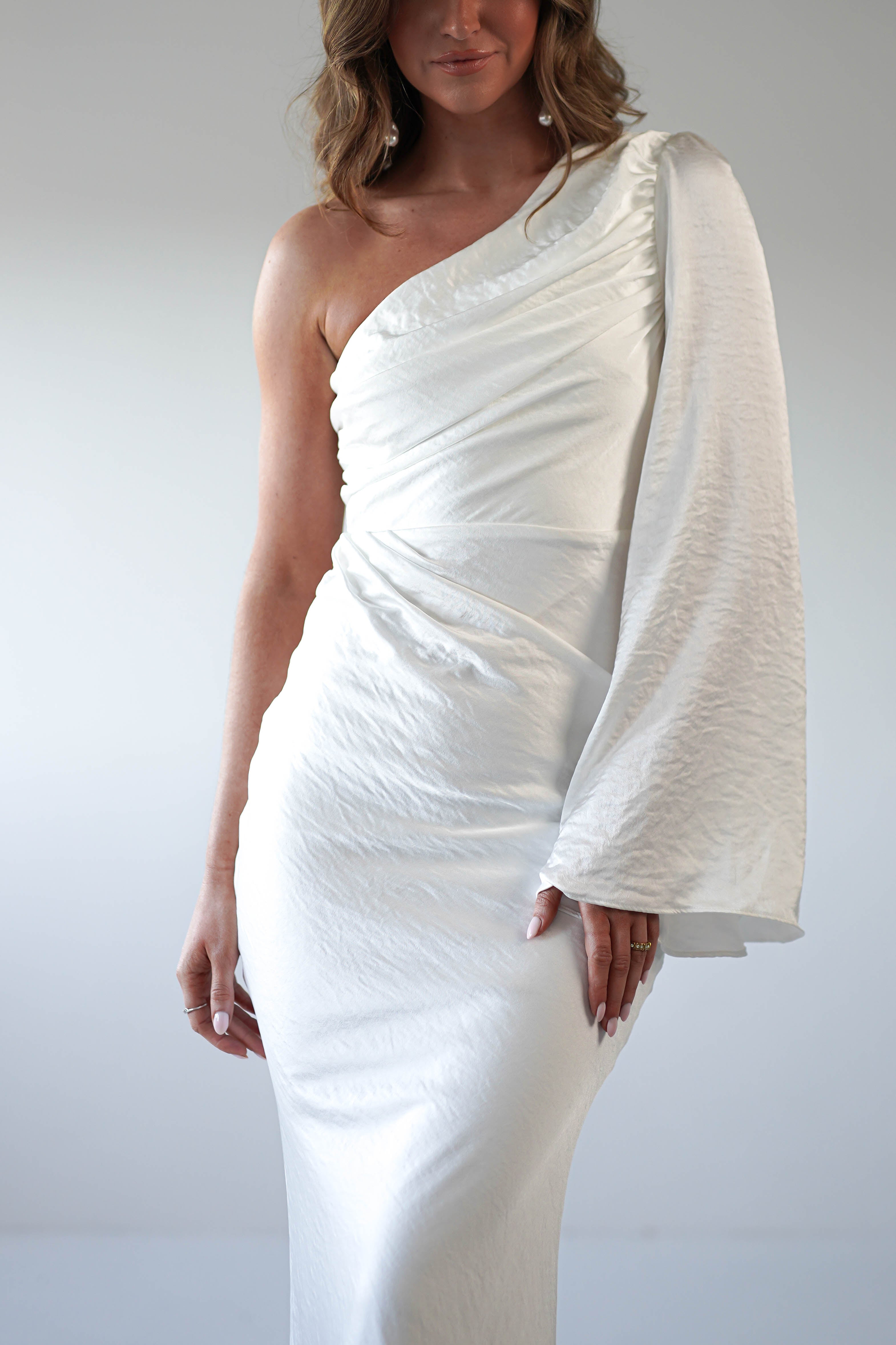 A woman wearing a fitted, formal dress with an asymmetrical neckline and a single long sleeve stands against a plain light background. The white dress features soft, ruched detailing and a smooth, elegant texture.
