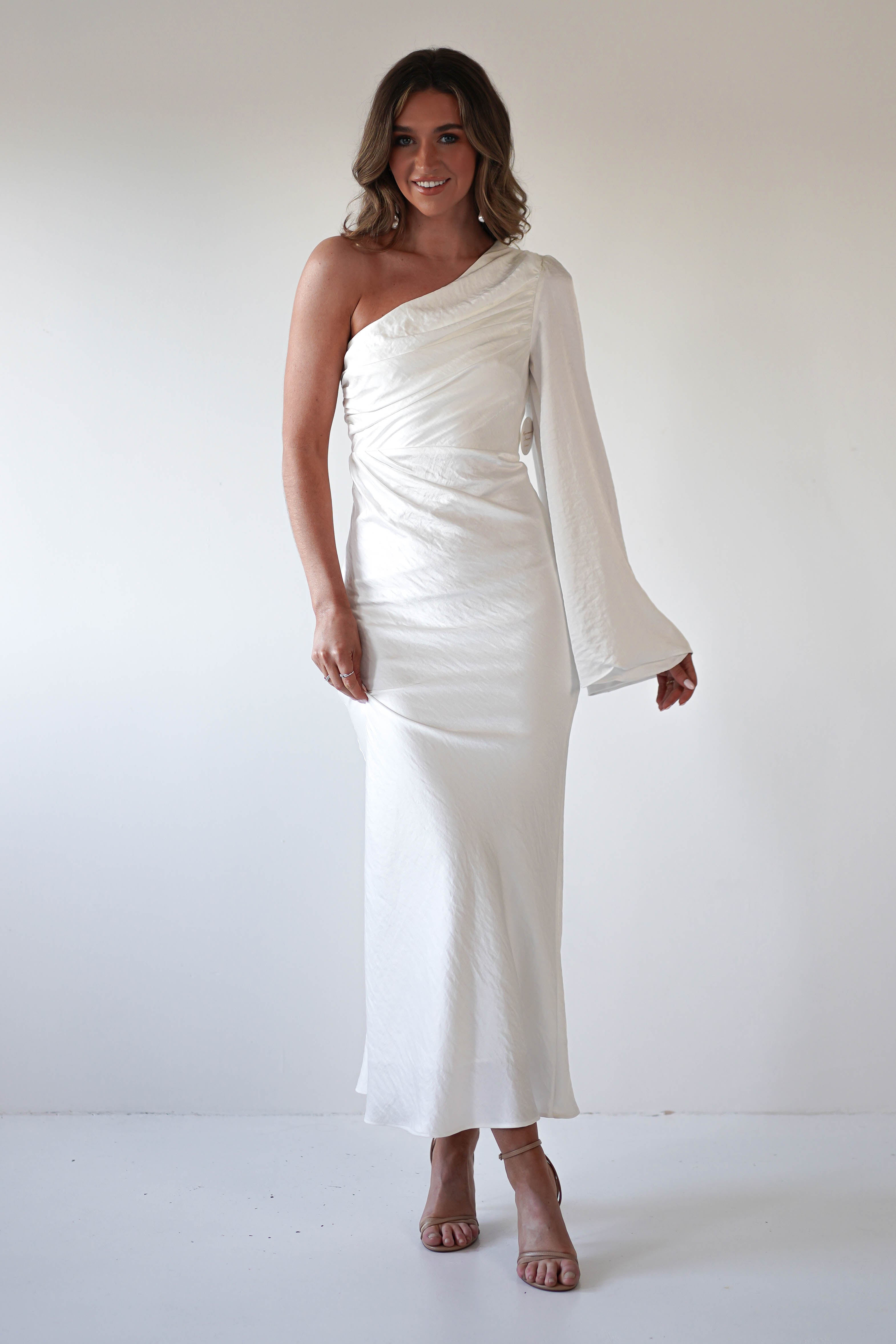 A woman in a formal dress with an asymmetrical neckline stands against a plain white background, smiling and posing with one arm slightly bent. She wears strappy nude heels and her hair is styled in loose waves.