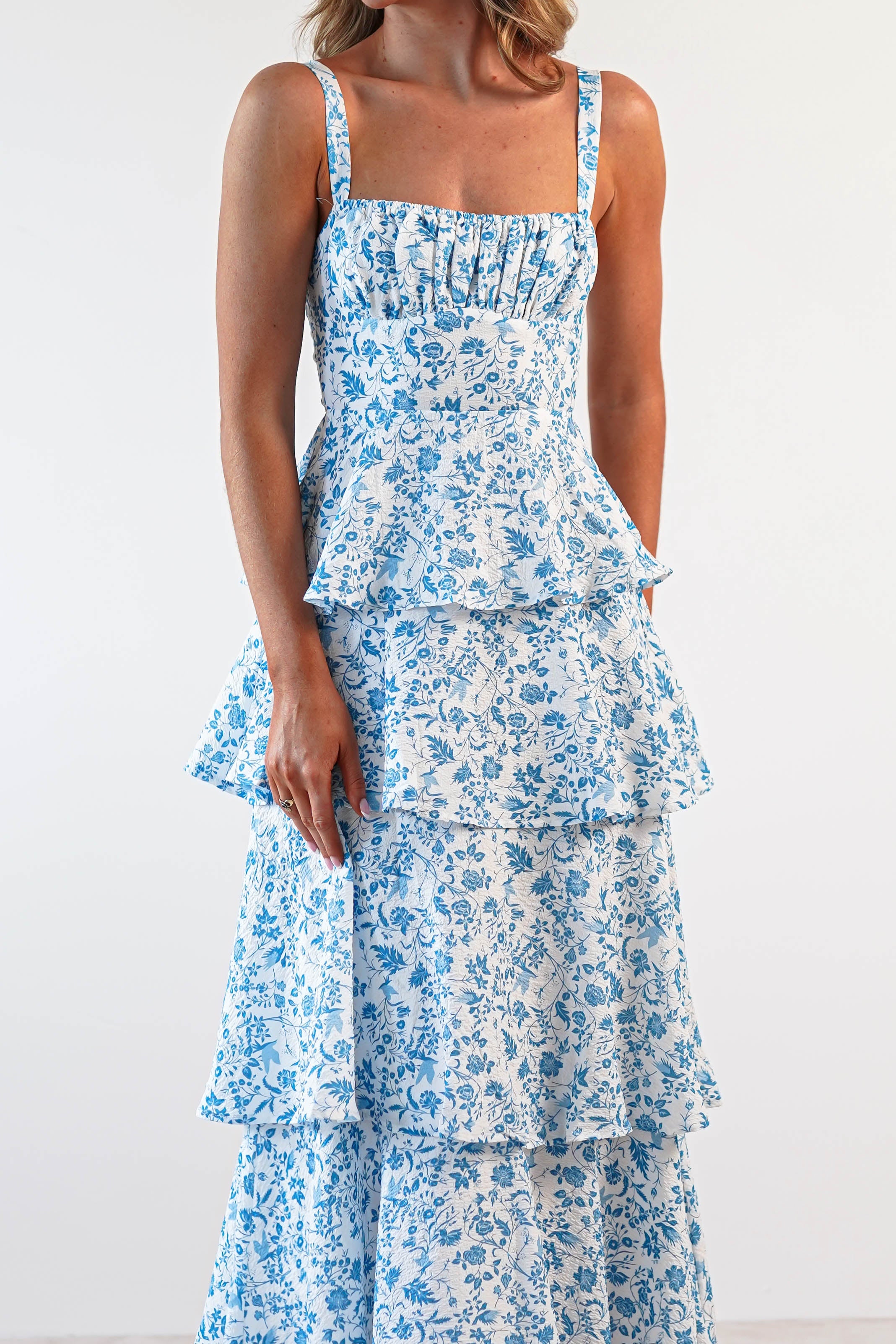 A woman in a sleeveless, tiered, blue-and-white floral dress stands against a plain background. The dress features a fitted bodice with ruffle detailing and tiered layers. Perfect for wedding guests or summer outings, her hair is down and her hands rest by her sides.