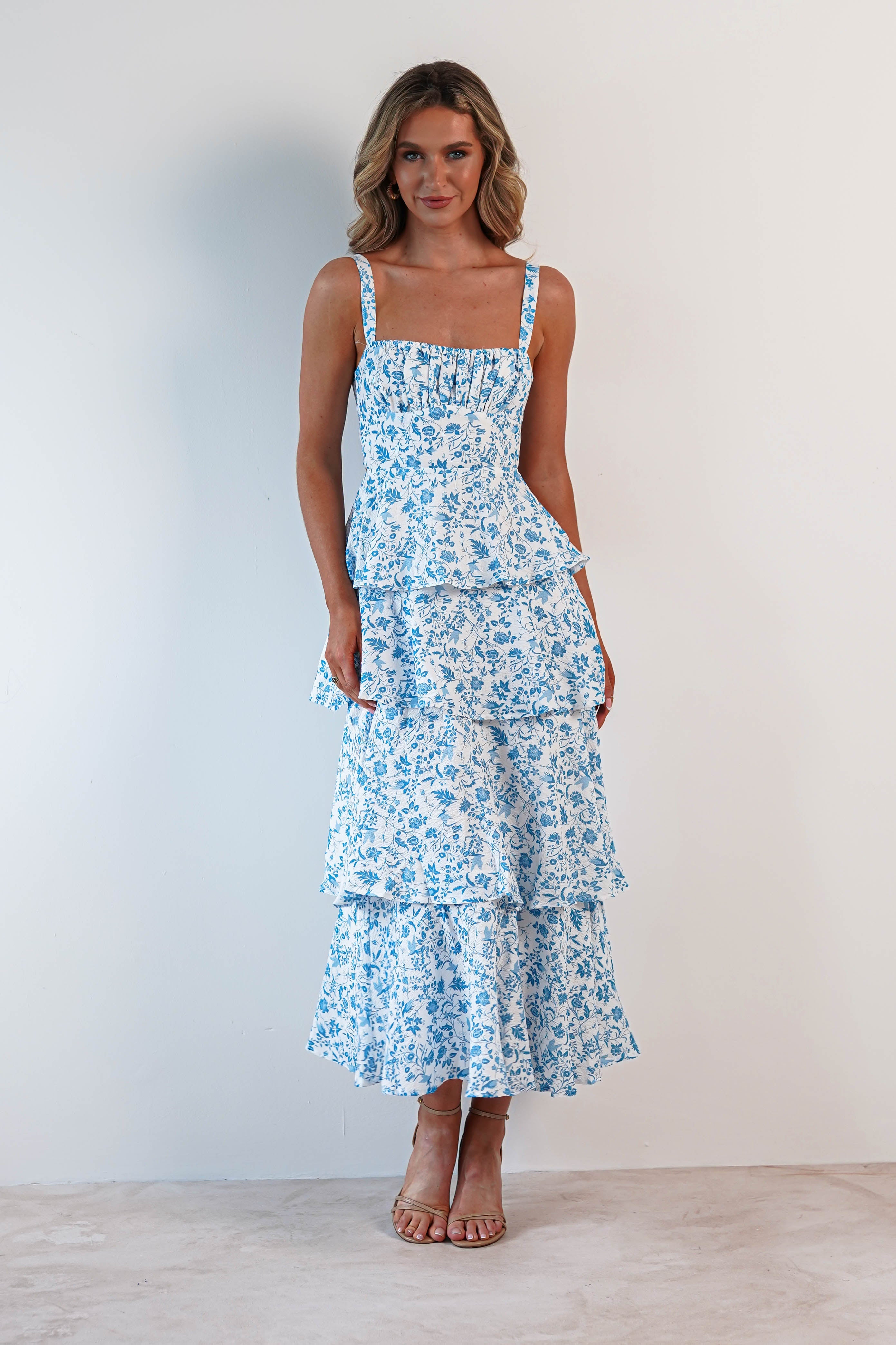 A woman stands with hands at her sides, showcasing a blue and white floral tiered dress with ruffle detail. The summer print maxi dress features a fitted bodice that flows into a full-length, ruffled skirt. Ideal for wedding guests, she pairs it with neutral open-toed heels against a light backdrop.