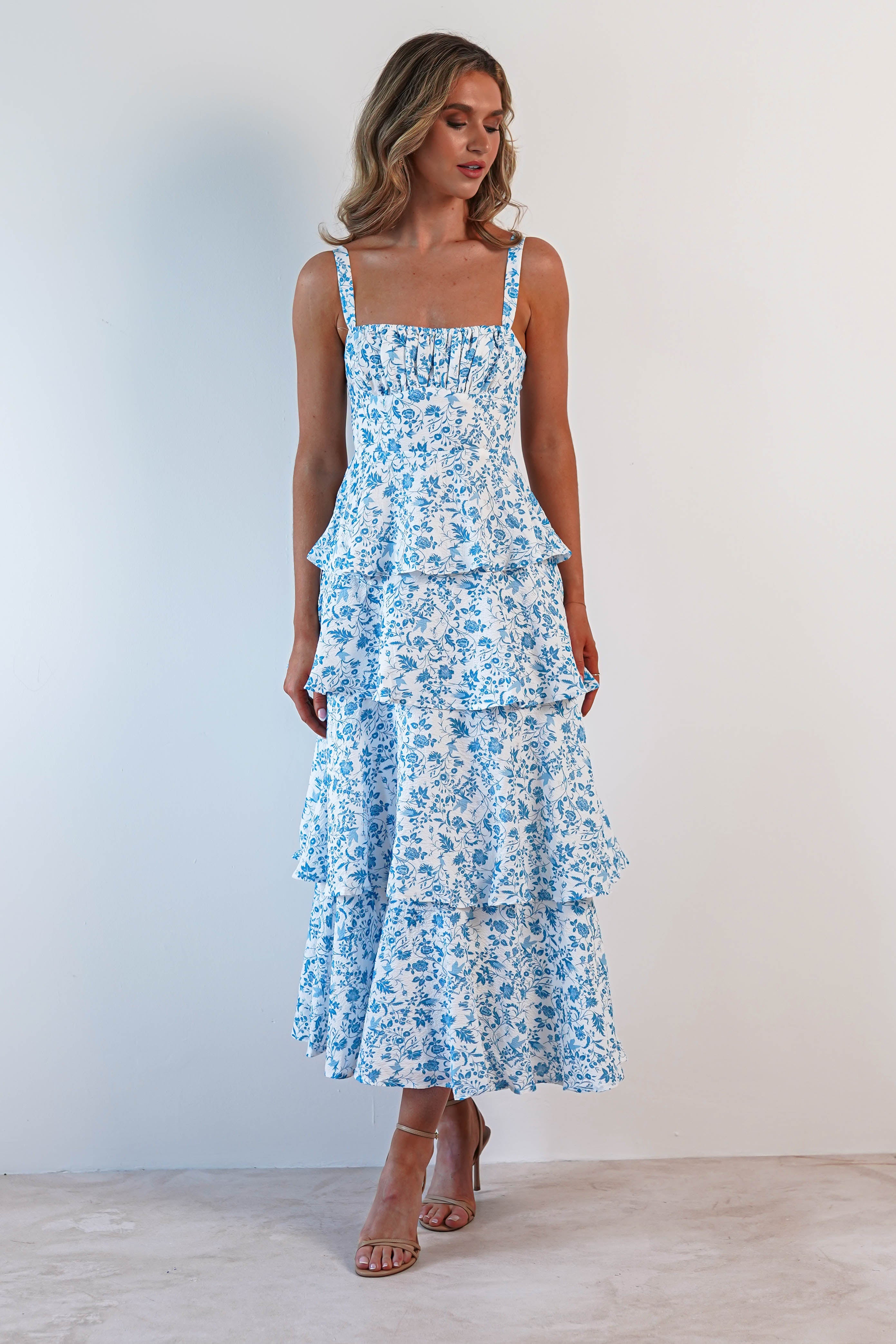 A woman in a long, summer print maxi dress with ruffle detail poses against a plain, light-colored background. Her light brown hair flows in loose waves, and she complements her look with strappy nude sandals, perfect attire for wedding guests.