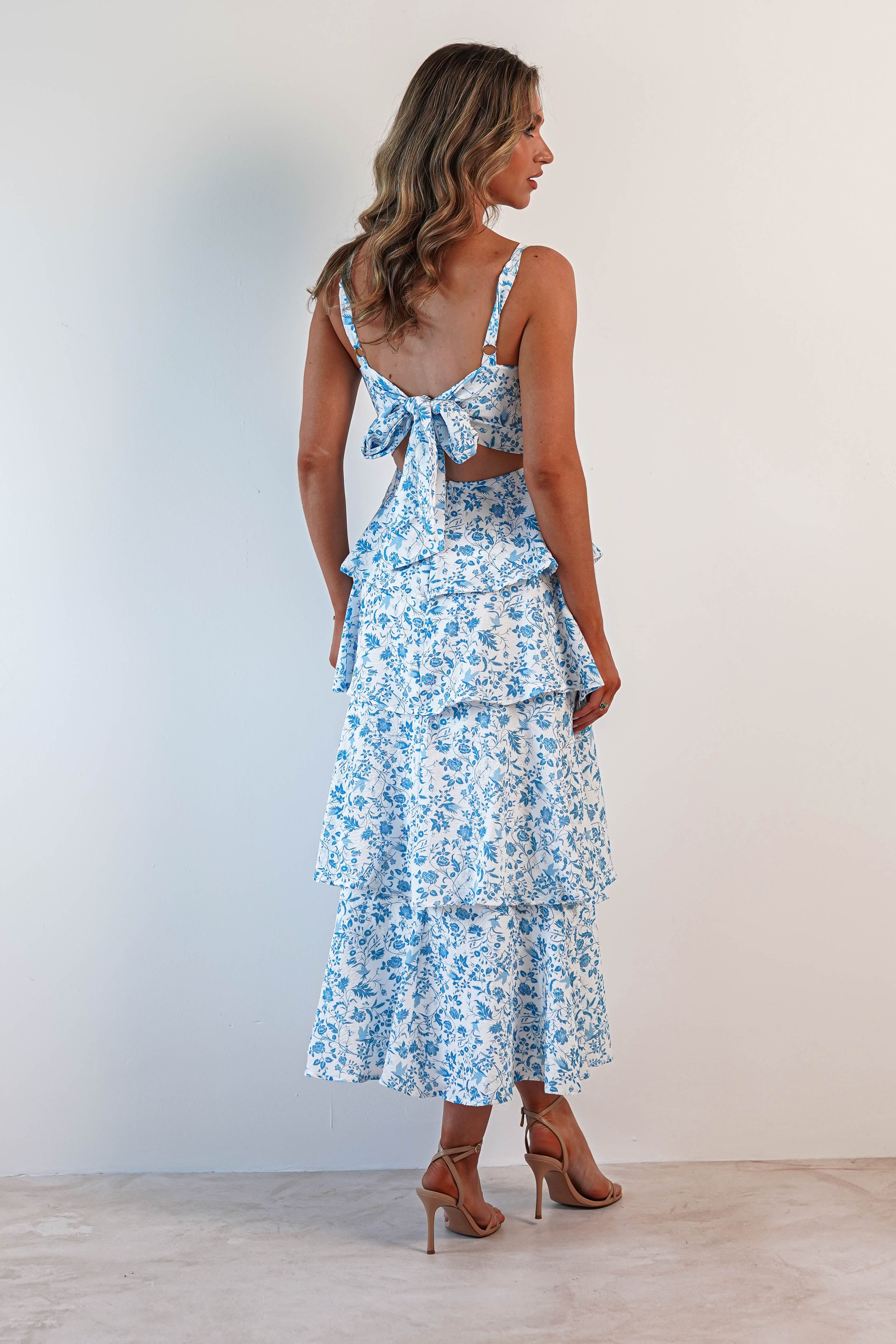 A woman stands sideways, showcasing a summer print maxi dress with blue and white florals, tiered ruffle details, and a bow at the back—perfect for wedding guests. Her long wavy hair complements her high-heeled sandals against a plain light background.