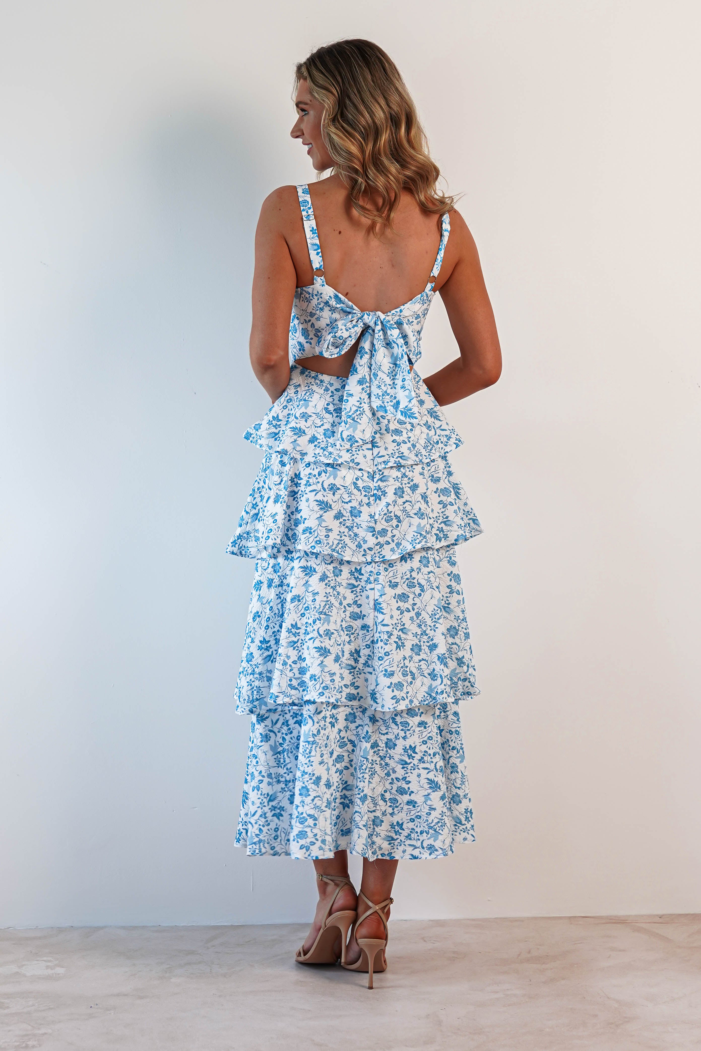 A woman stands facing a wall, wearing a sleeveless maxi dress adorned with blue floral patterns on a white background. The summer print features ruffle details and a large bow at the back. She is wearing strappy heels, her hair styled in loose waves—a perfect look for wedding guests.