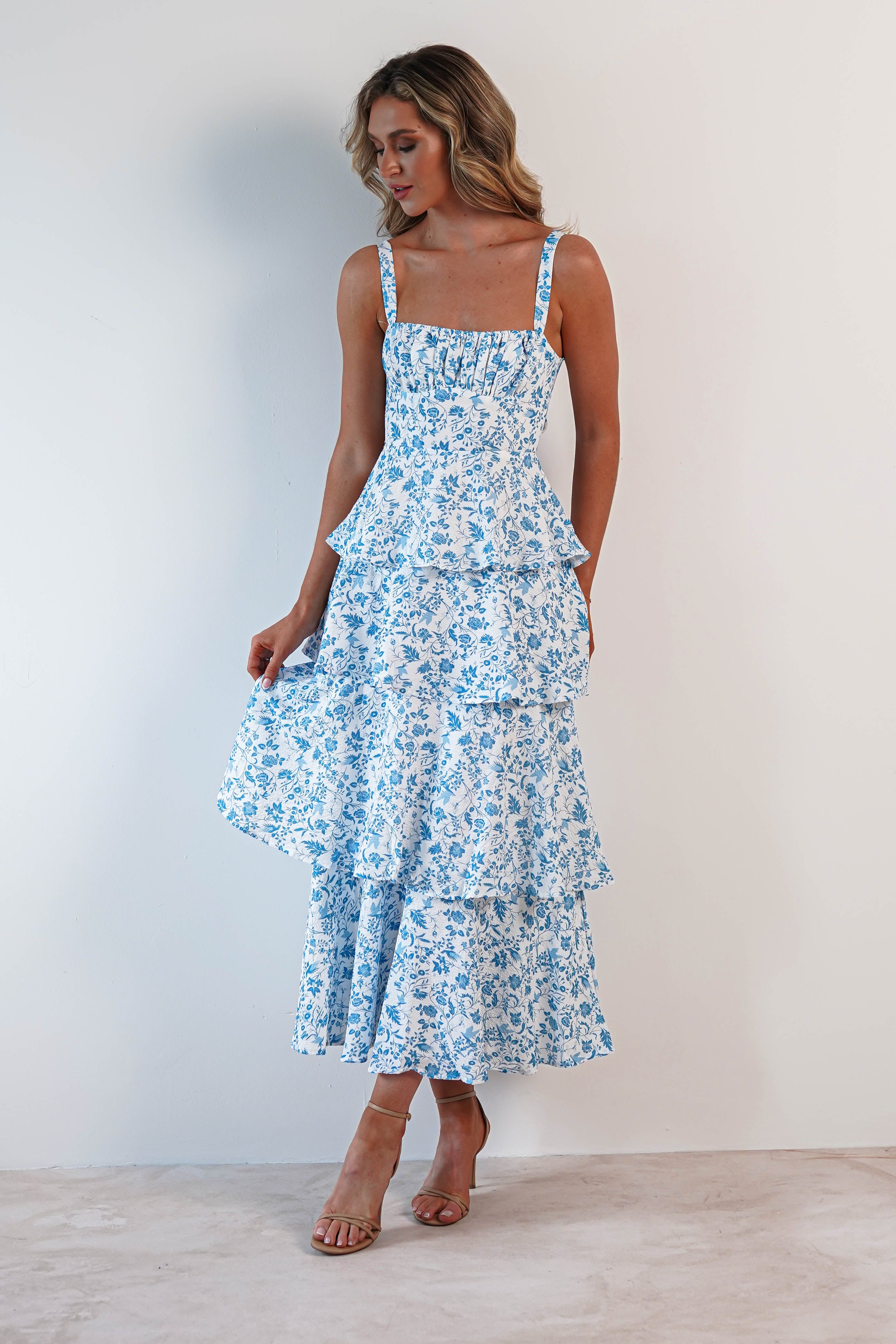 A woman in a blue and white floral tiered dress with ruffle detail stands against a plain white background. The dress features a square neckline and spaghetti straps. Perfect for wedding guests, she pairs it with nude heels, her hair styled in loose waves.