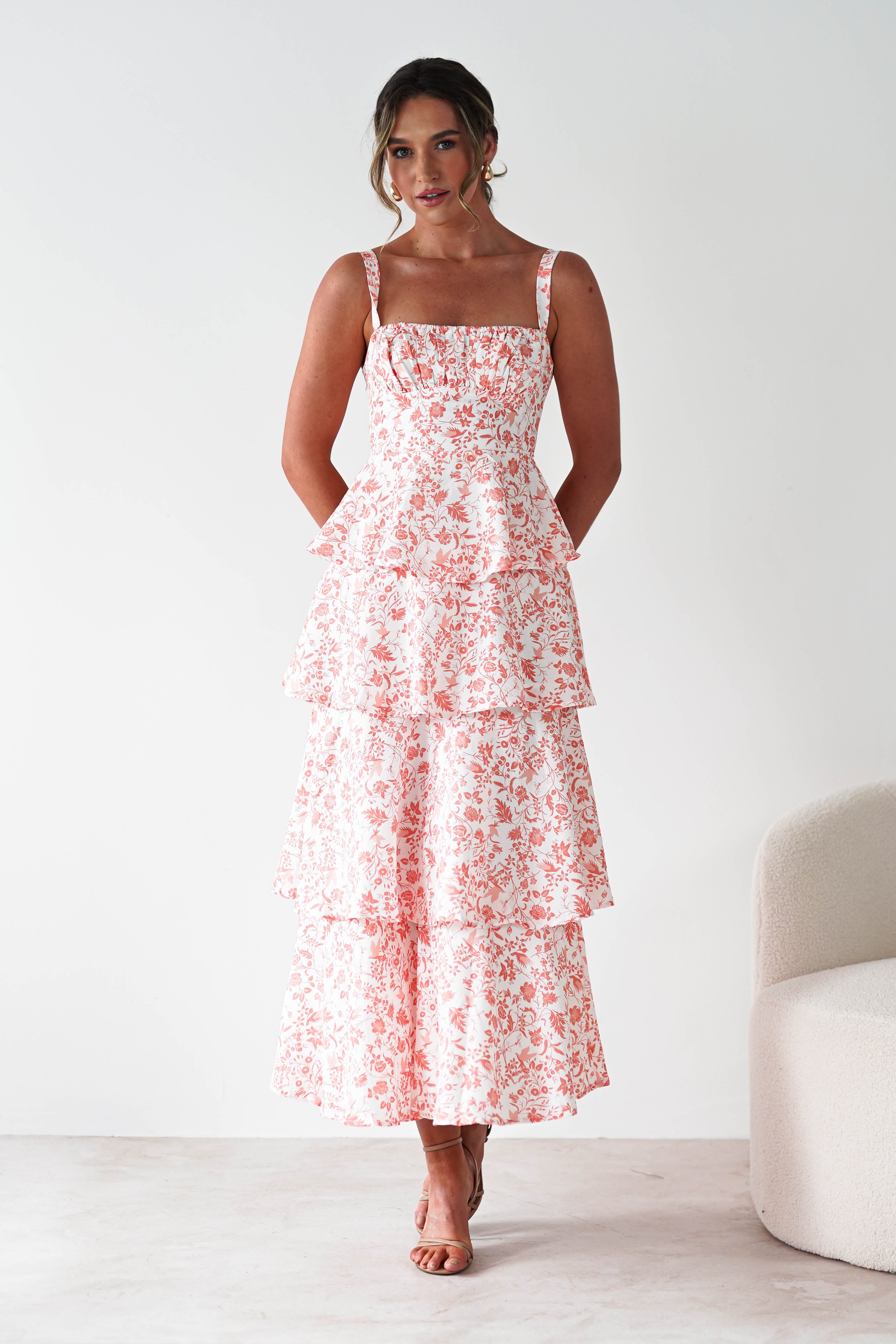 A woman stands indoors wearing a sleeveless, tiered, ankle-length white dress with a pink floral pattern and ruffle detail. She faces forward with her hands behind her back. A light-colored chair is partially visible on the right—perfect for wedding guests.