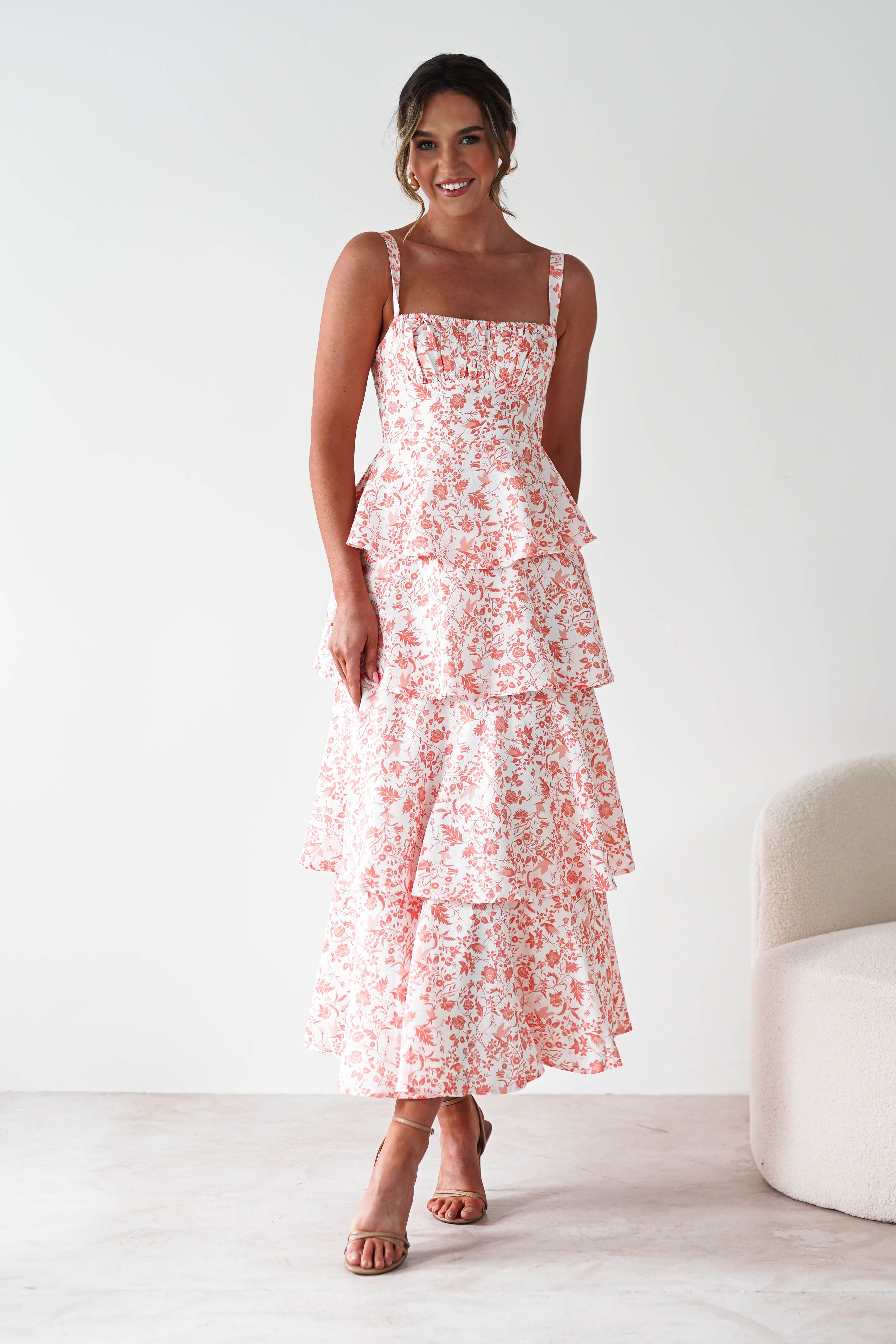 A woman stands smiling in a sleeveless, tiered maxi dress with pink floral and ruffle detail. She wears strappy heels—an ideal look for wedding guests—posing in a light, minimal room with a curved beige chair.