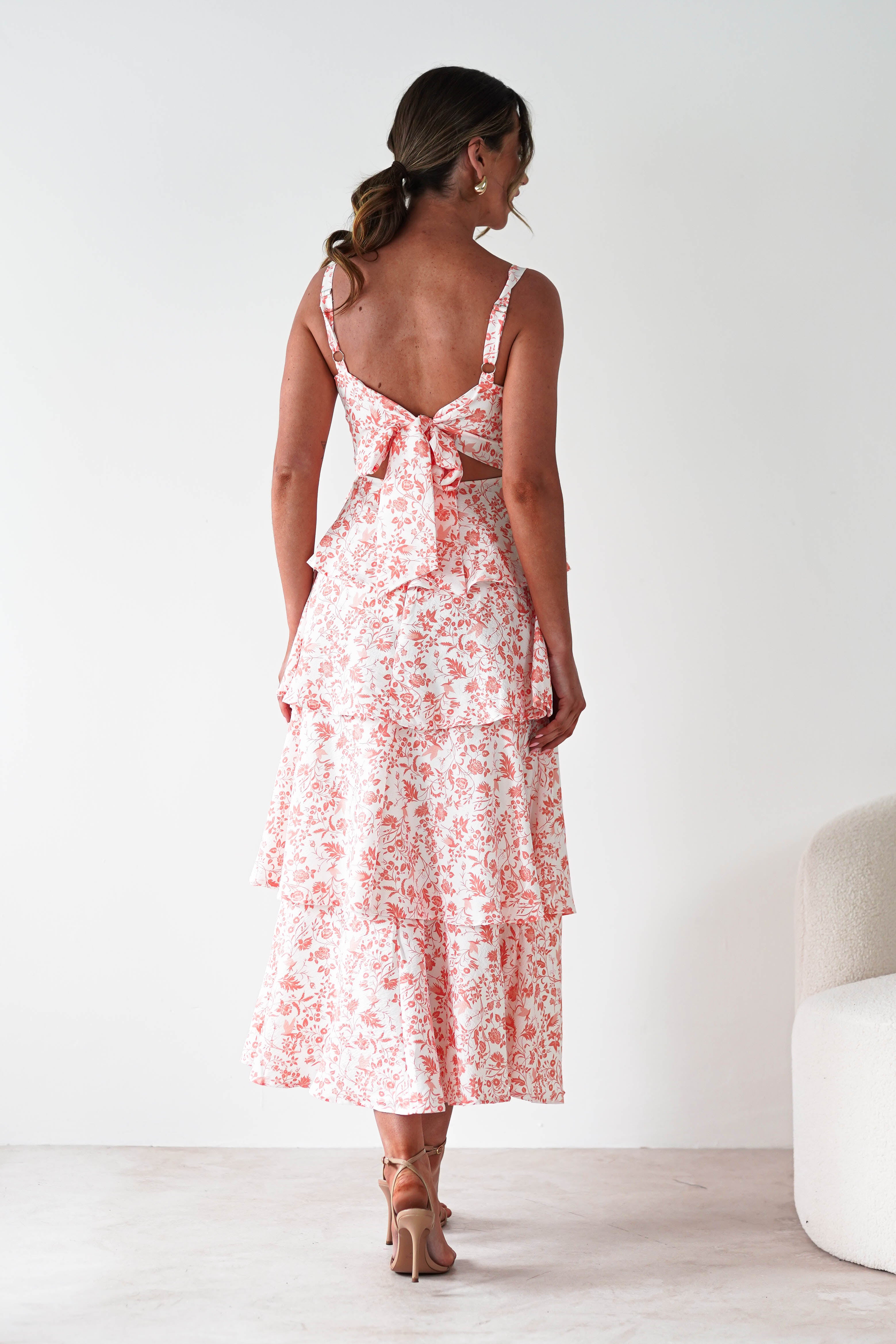 A woman in a summer orange print maxi dress with ruffle detail stands facing away in a minimalist room with white walls and a light-colored chair—an elegant choice for wedding guests.