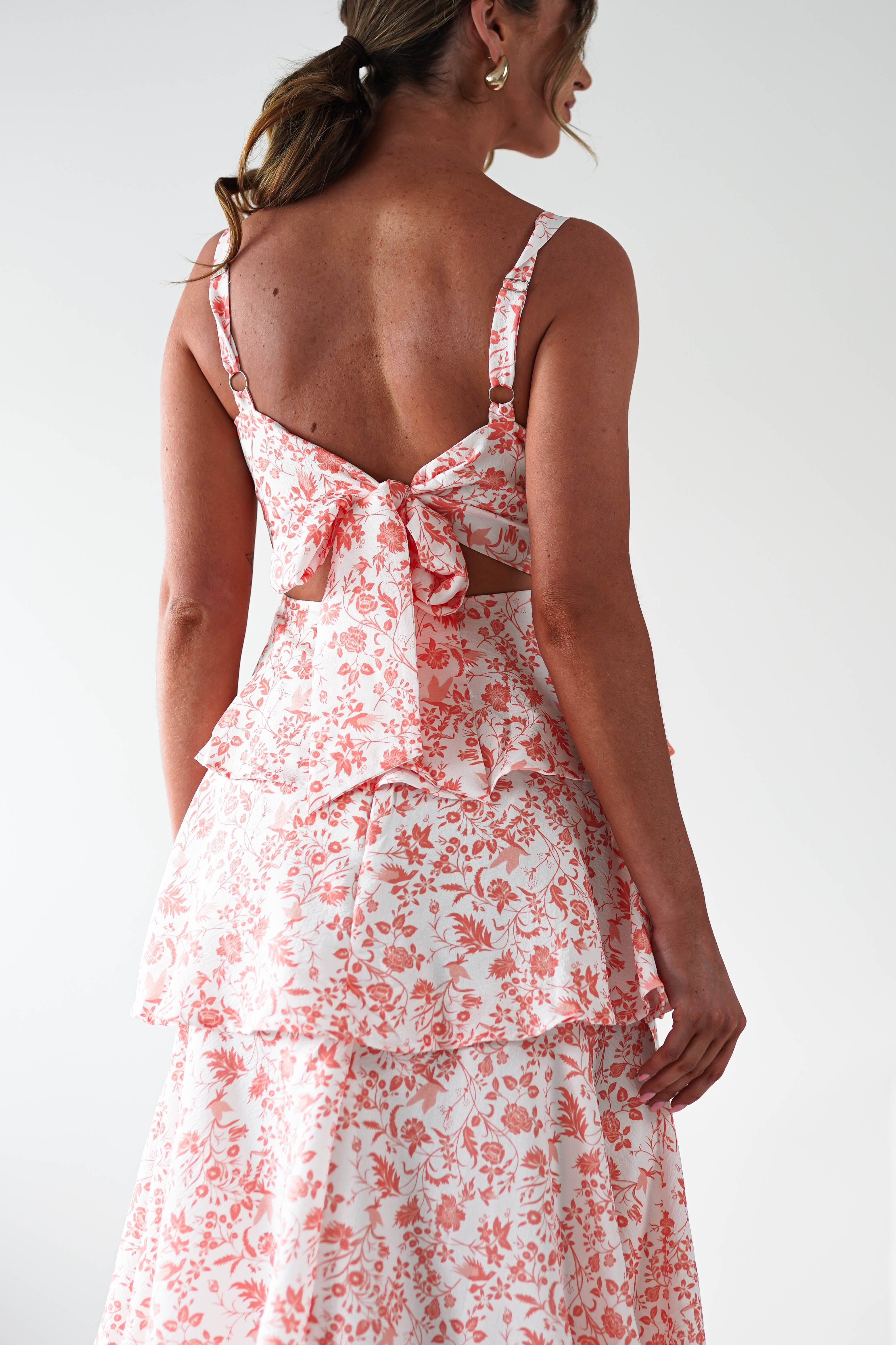 A woman stands with her back to the camera, wearing a sleeveless dress with pink floral patterns and ruffle detail, perfect for wedding guests. The dress features ruffled layers, a large bow at the back, and her brown hair in a loose ponytail.