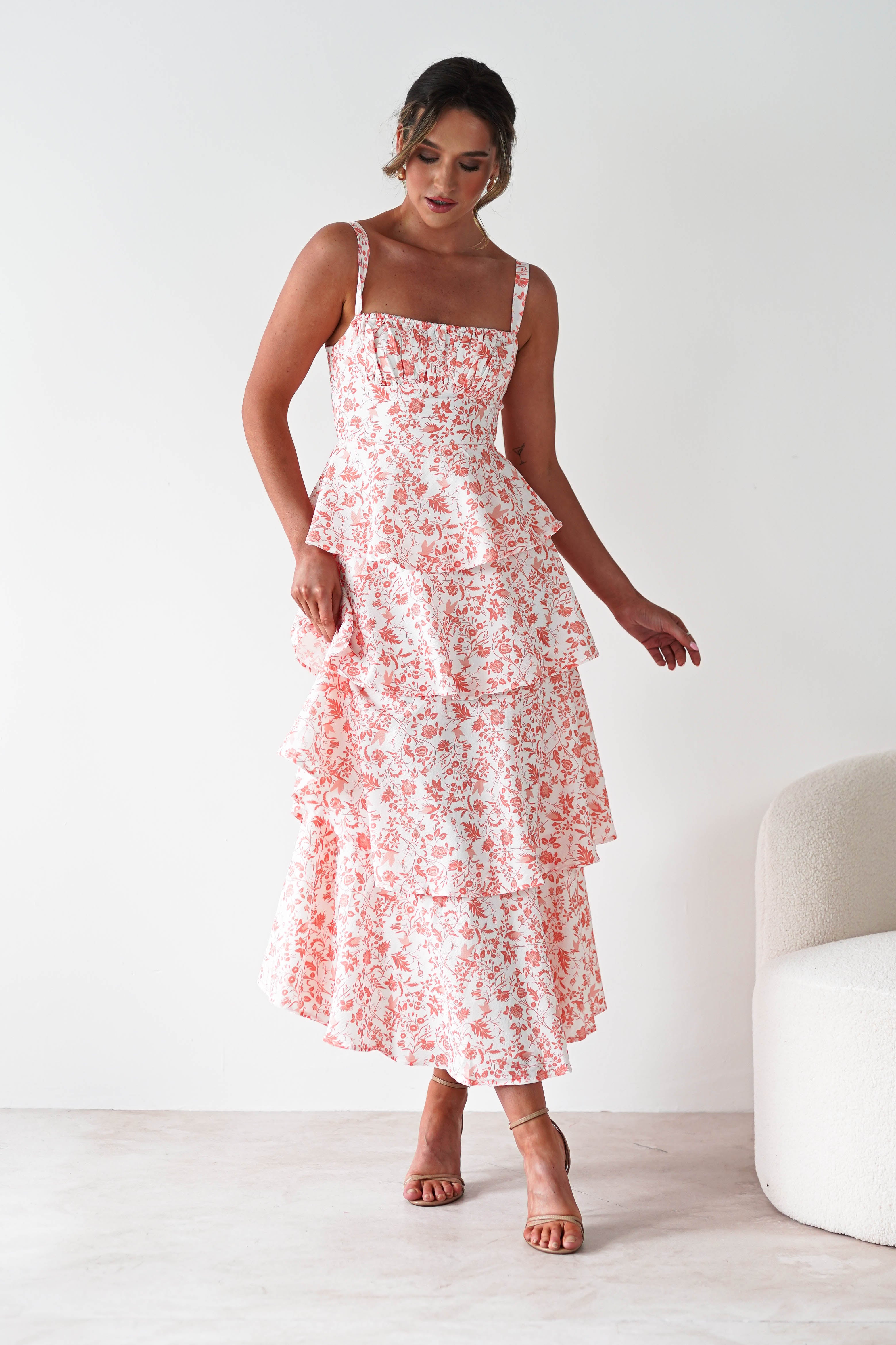 A woman wearing a sleeveless, tiered, pink and white floral dress with ruffle detail stands indoors near a white chair, looking down with one hand slightly raised.